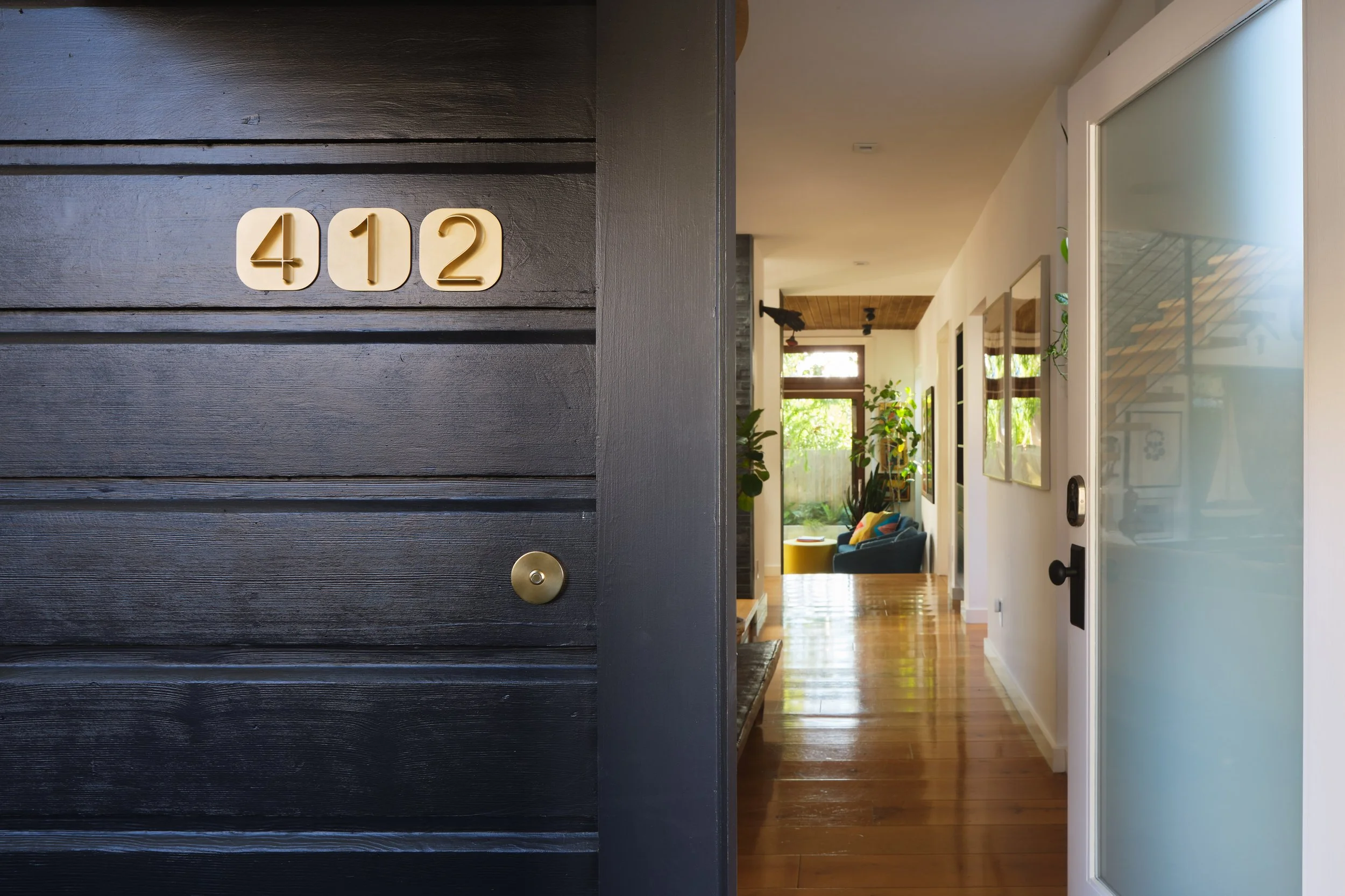 House number 412 on a dark wooden door, with a view into a bright hallway with hardwood flooring, potted plants, colorful furniture, and windows letting in natural light.