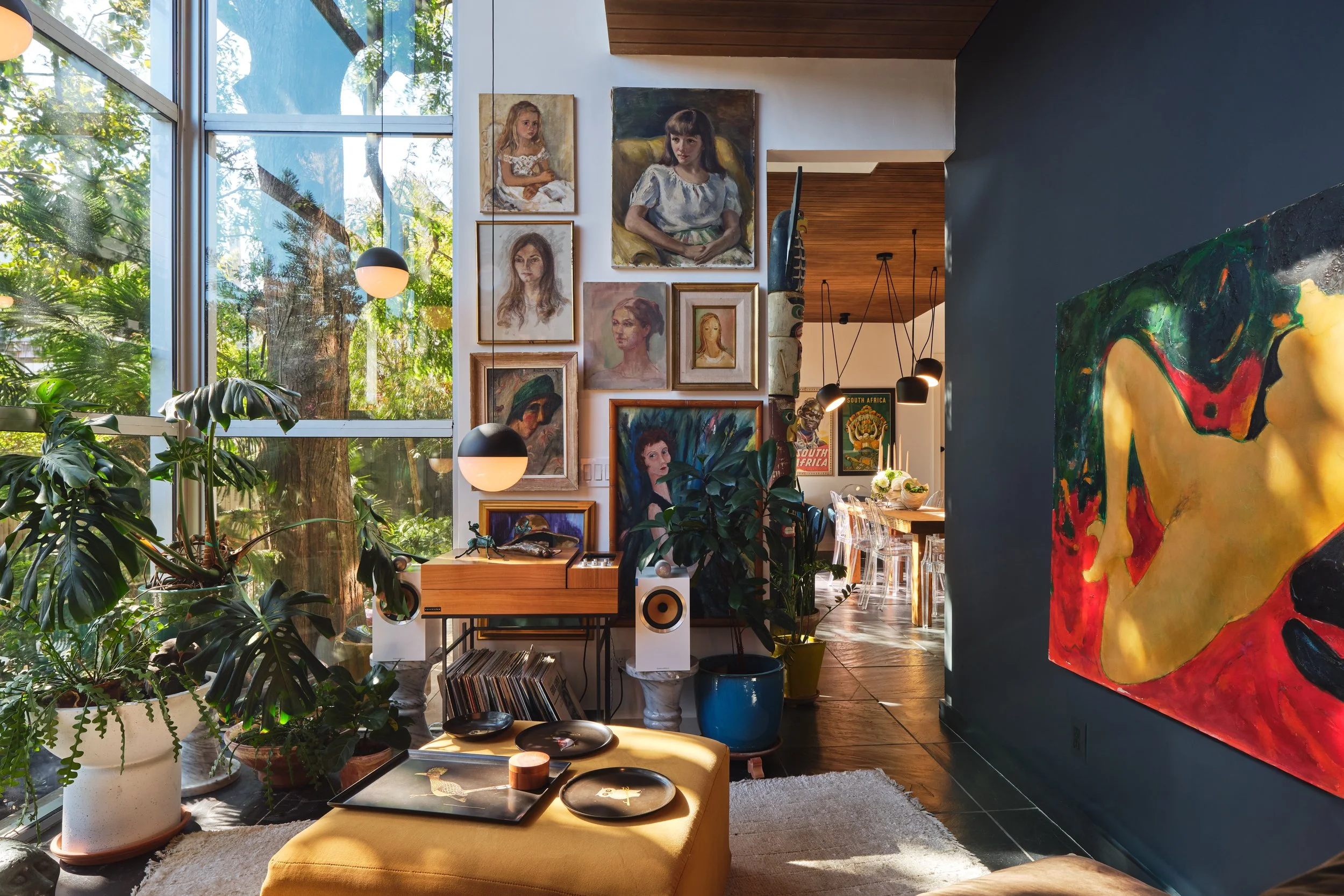 Interior of an art-filled living space with large windows, green plants, framed paintings on the wall, and a yellow ottoman with decorative plates.