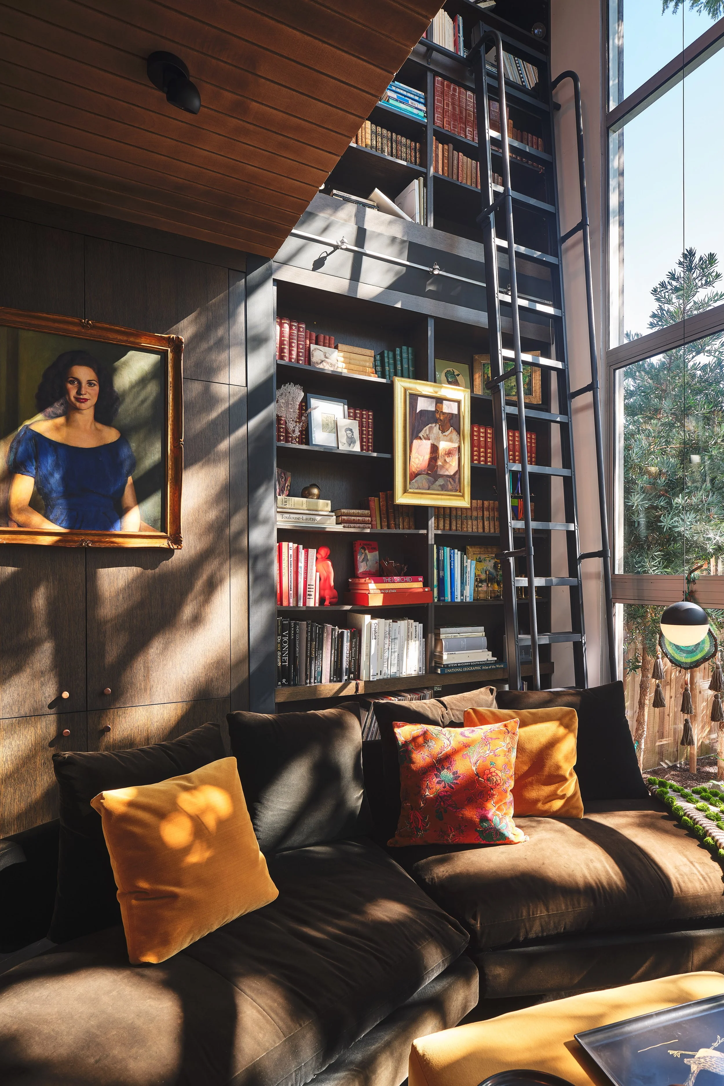 A cozy living room with a large black leather sectional sofa adorned with yellow and patterned pillows. Behind the sofa, a tall, black bookshelf filled with books and framed photos, with a ladder attached for reaching higher shelves. To the side, a large window lets in natural light, with a view of greenery outside. The room features warm lighting, a wooden ceiling, and a hanging spherical lamp.