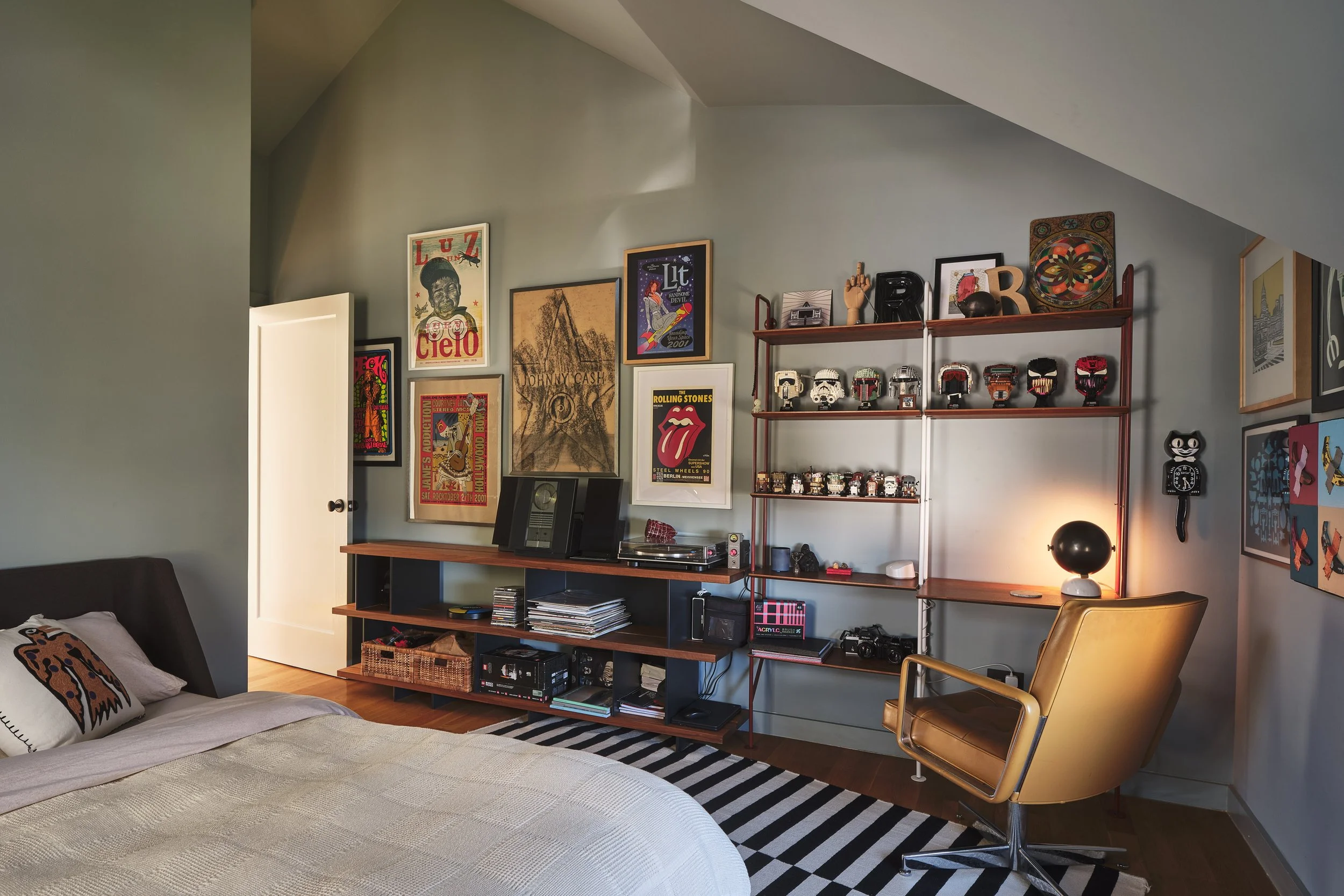 A cozy living room corner with a bed, pillows, and a gallery wall with posters and framed pictures. There are shelves with decorations and collectibles, a lamp, and a golden chair.