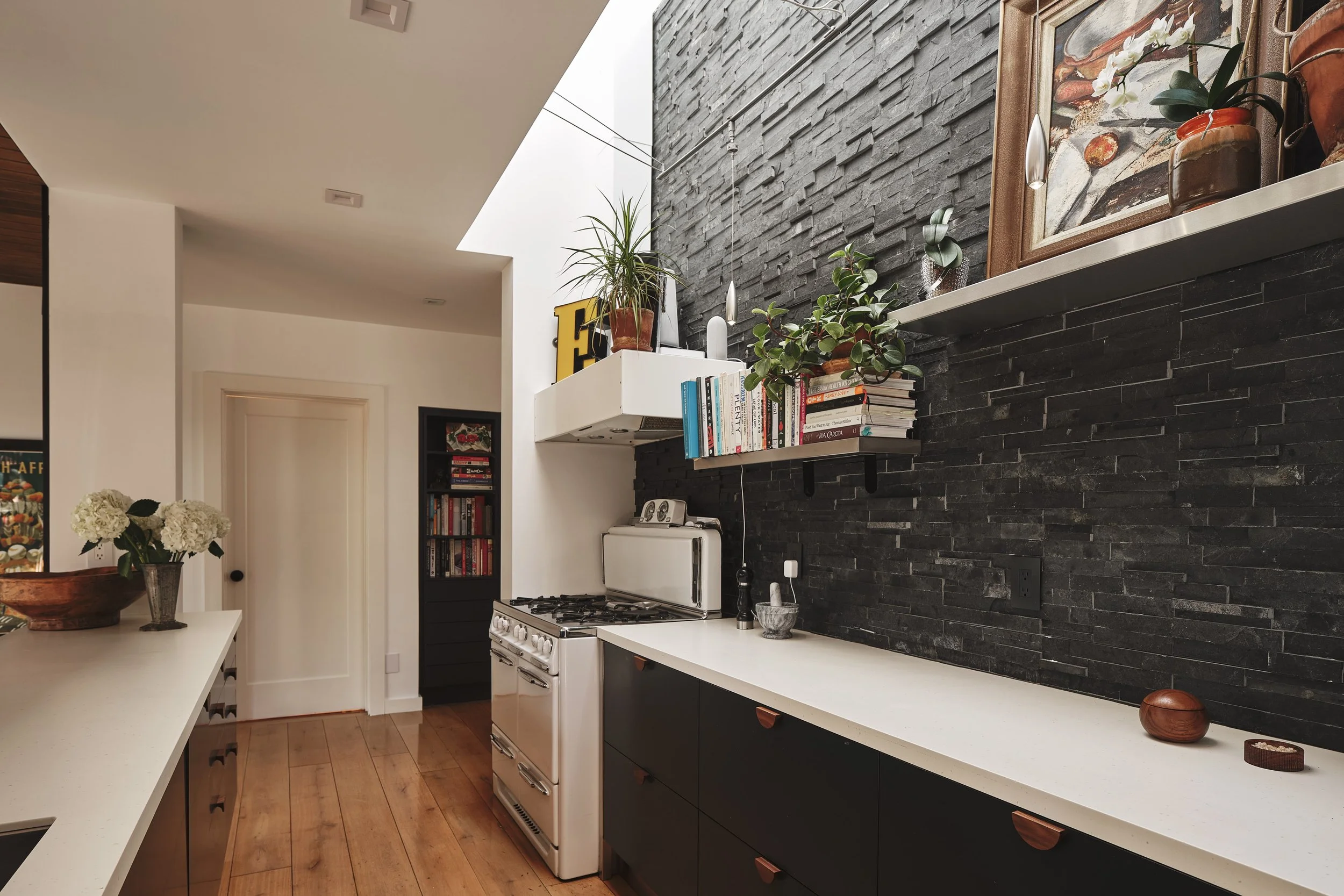 Modern kitchen with black stone accent wall, white countertops, a white stove, shelves with books and plants, and decorative vases.