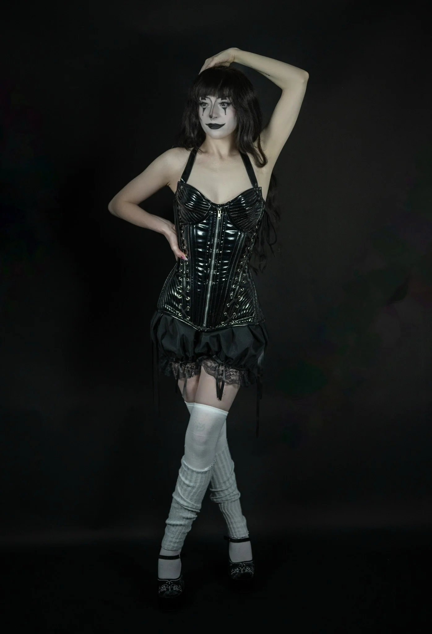 Woman in a black latex corset, black skirt, white thigh-high socks, and black shoes with lace, posing with one hand on her hip and the other raised above her head against a black background. She has dark makeup and long dark hair with bangs.