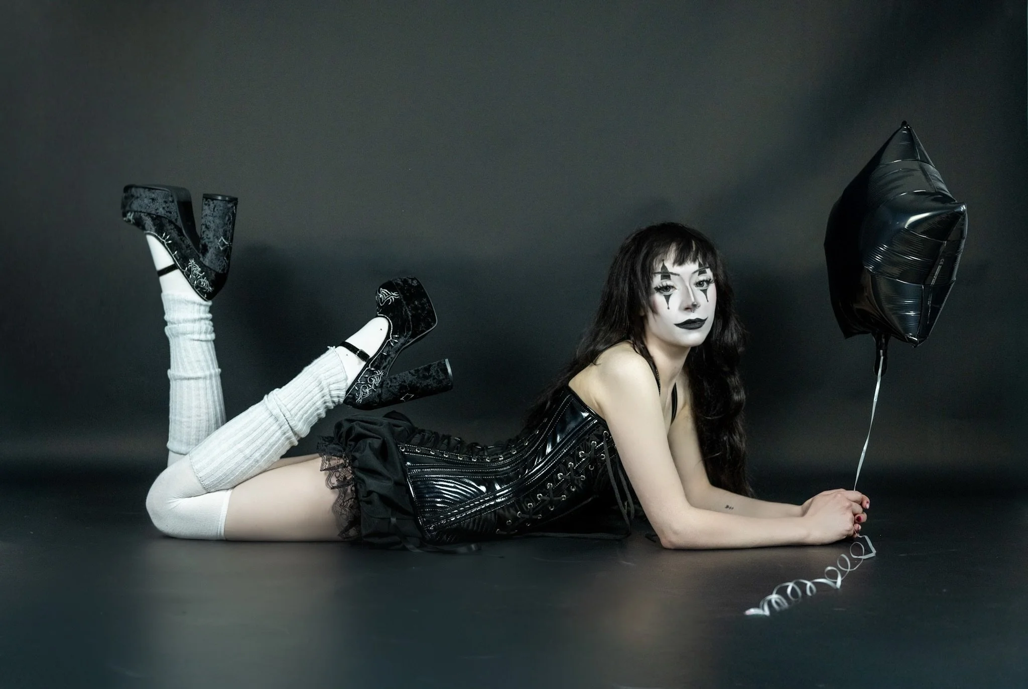 A woman with dark hair and dramatic makeup is lying on her stomach against a dark background. She is wearing a black, shiny, striped dress with a lace trim, white thigh-high stockings, and black high heels. She is holding a balloon and has a curling ribbon attached to it.