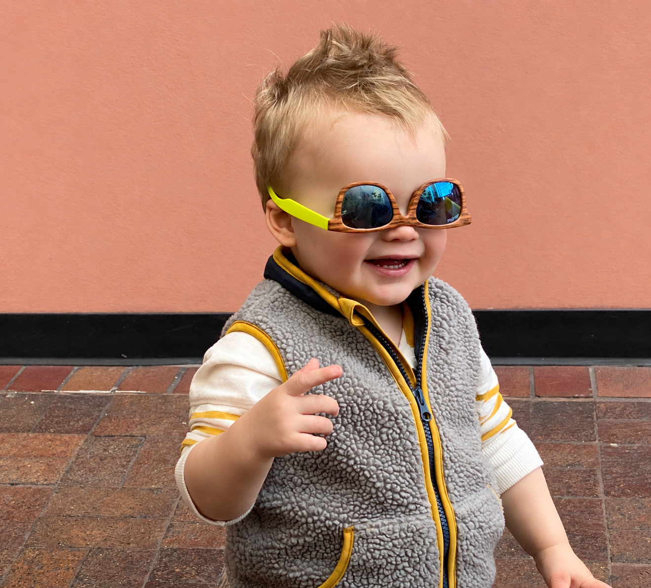 A young child with blond hair wearing sunglasses with reflective blue lenses, a patterned vest, and a striped long sleeve shirt, smiling and pointing with their finger, standing against a pink wall on a brick sidewalk.