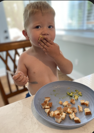 A young shirtless boy with blonde hair eating a piece of bread or toast at a kitchen table. There is a blue plate with bread crumbs and small pieces of bread in front of him.