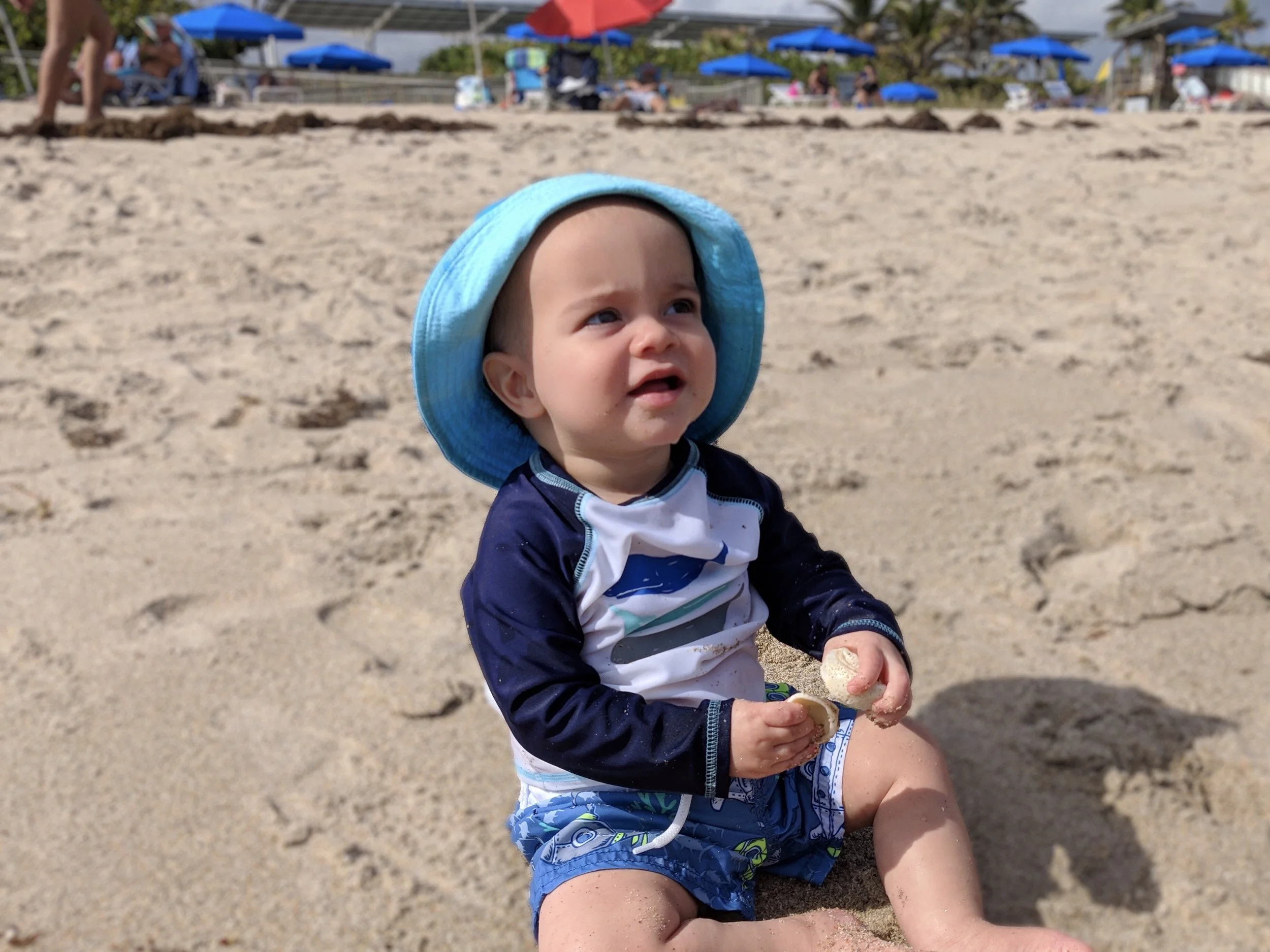 A young boy wearing a blue sun hat and swimwear sits on the sandy beach, holding an ice cream cone with a bite taken out of it, with beach umbrellas and people in the background.