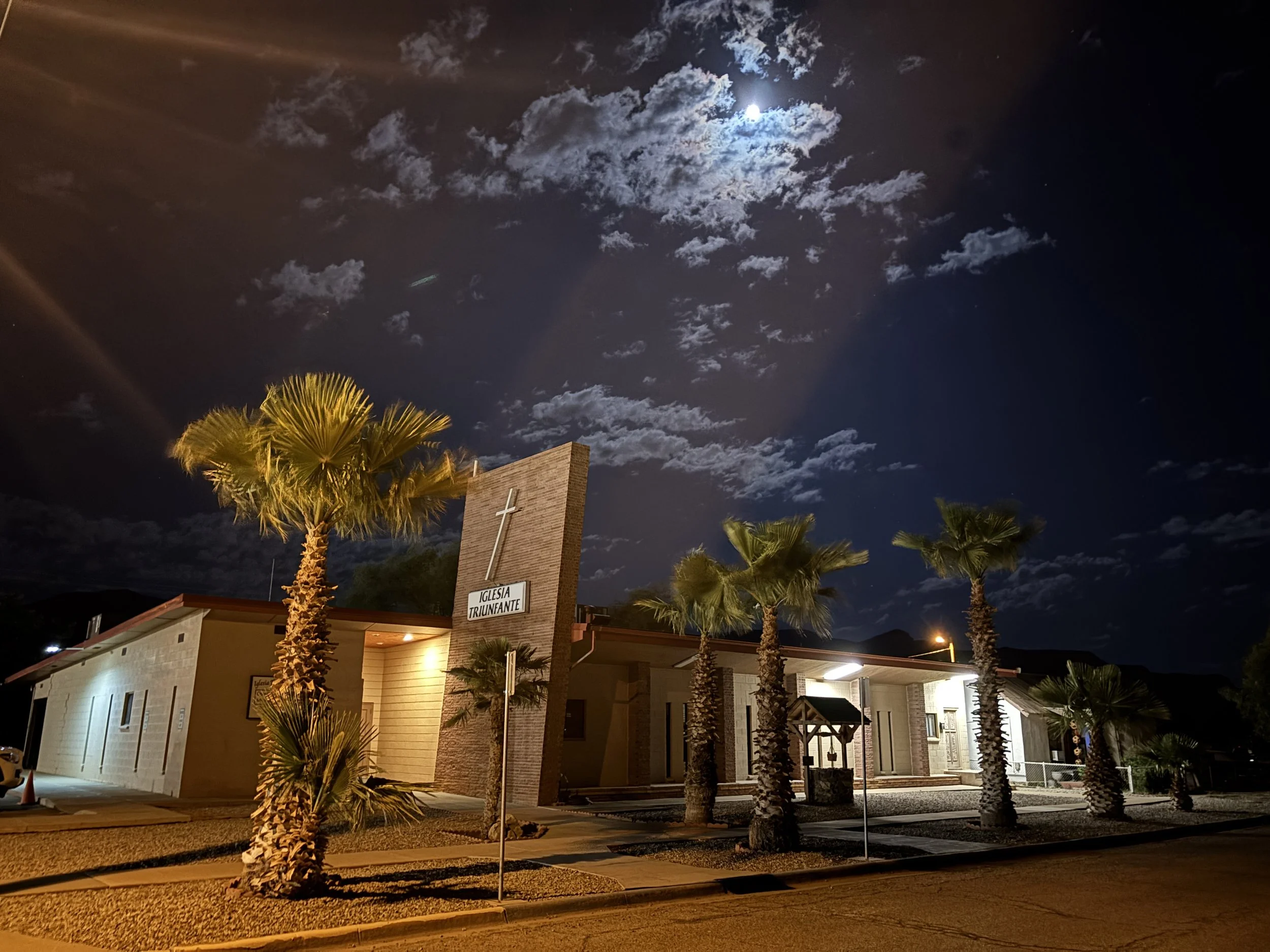 Una iglesia cristiana llamada 'Iglesia Triunfante' al atardecer, con árboles de palmeras y nubes en el cielo iluminado por la luna.