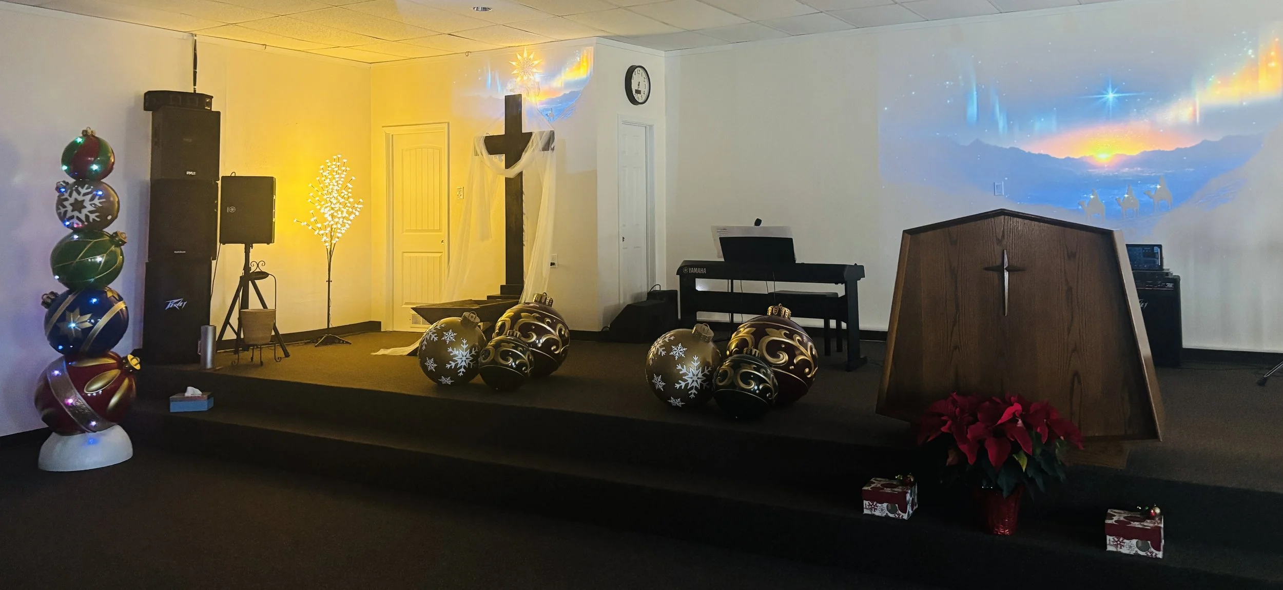 Escenario decorado para celebración navideña con regalos, bolas navideñas, árbol de Navidad con luces, proyector de escena navideña en la pared, crucifijo de madera, piano con monitor y otros equipos de sonido, flores de poinsettia, bohemia con puntas de color rojo y blanco, decoración festiva.