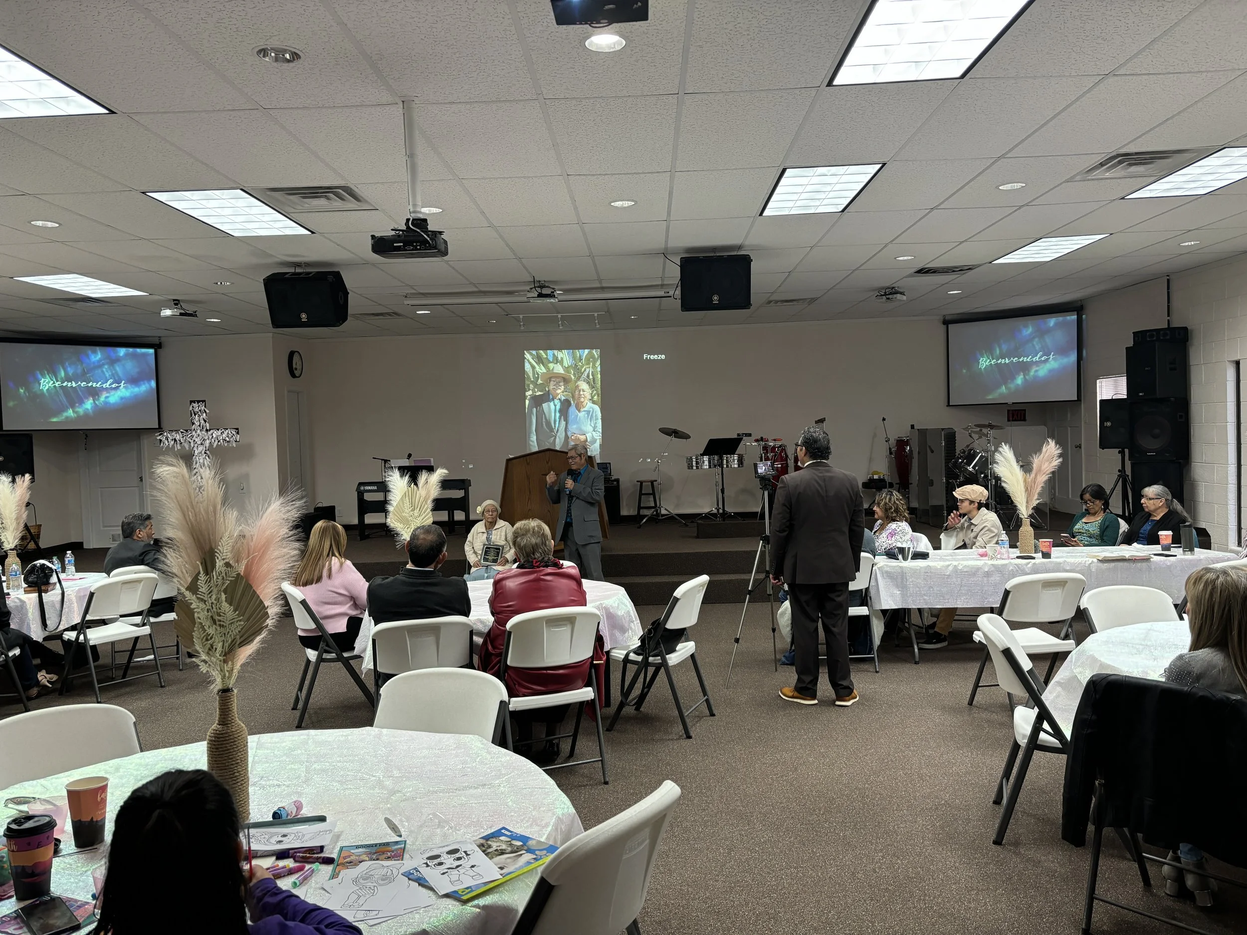 Sala de reuniones con varias personas sentadas alrededor de mesas, una persona de pie frente a la audiencia, pantallas en la pared exhiben palabras y una foto, decoraciones con plantas y cruces, ambiente de conferencia o servicio religioso.