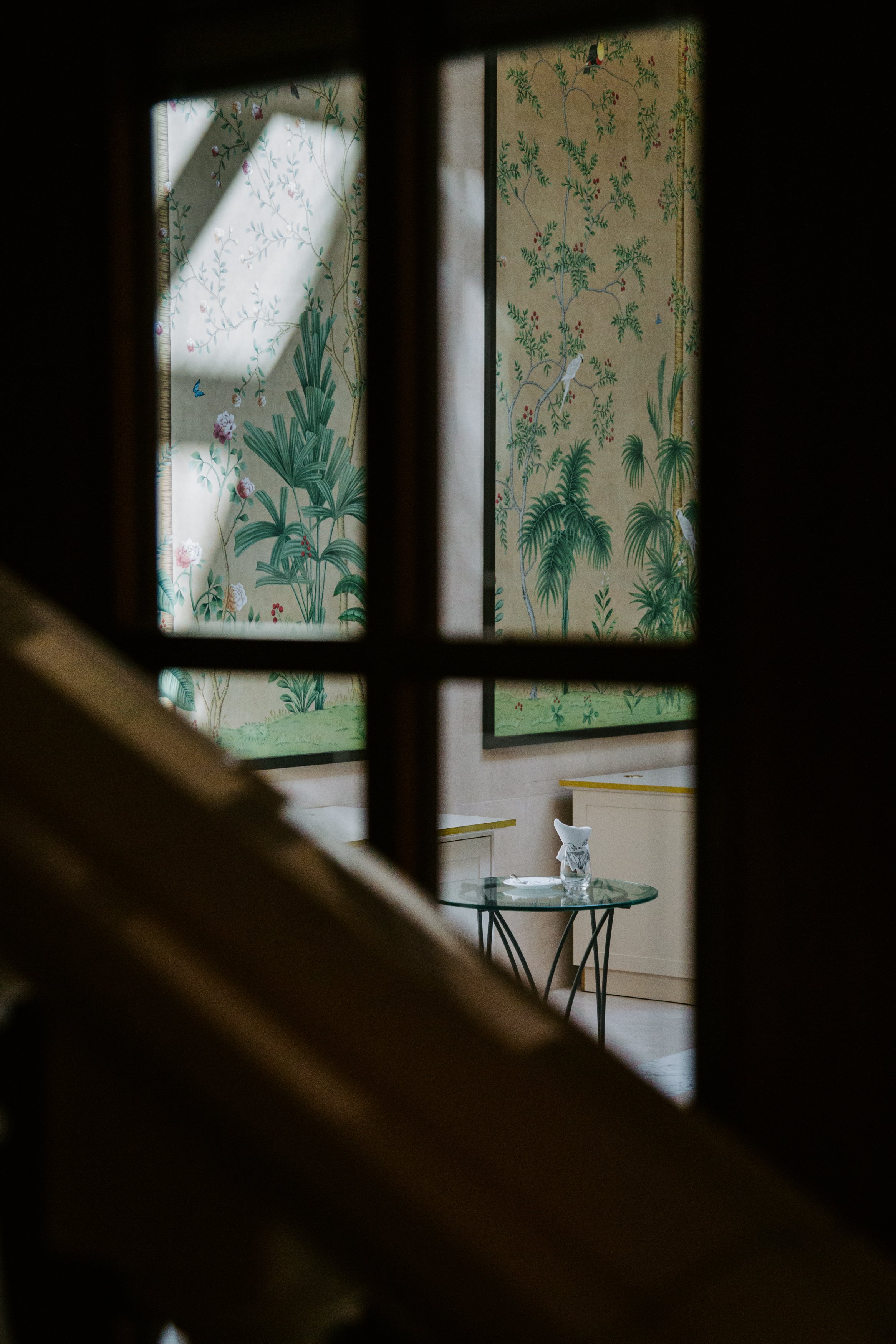 View of a room through a small window with wooden frame, showing a patterned wallpaper with green foliage and birds, a small white table with a glass pitcher and a folded napkin on top.