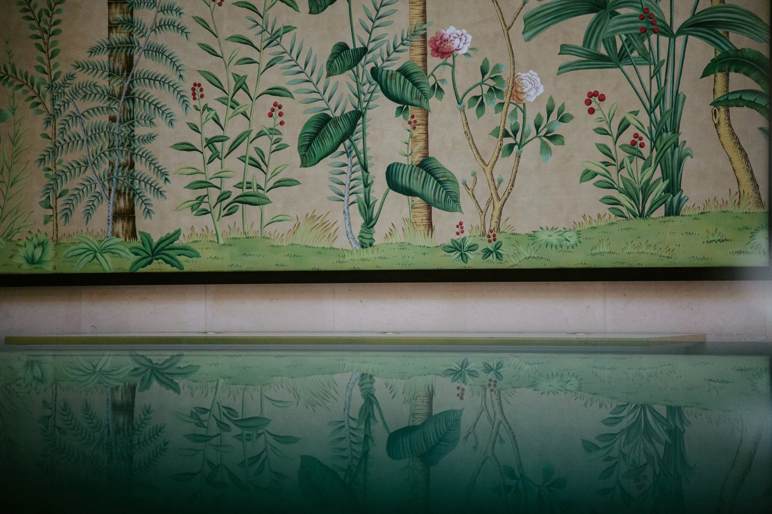 A detailed mural of lush green plants, trees, and flowers painted on a wall, with their reflection visible in a mirror or glass surface below.