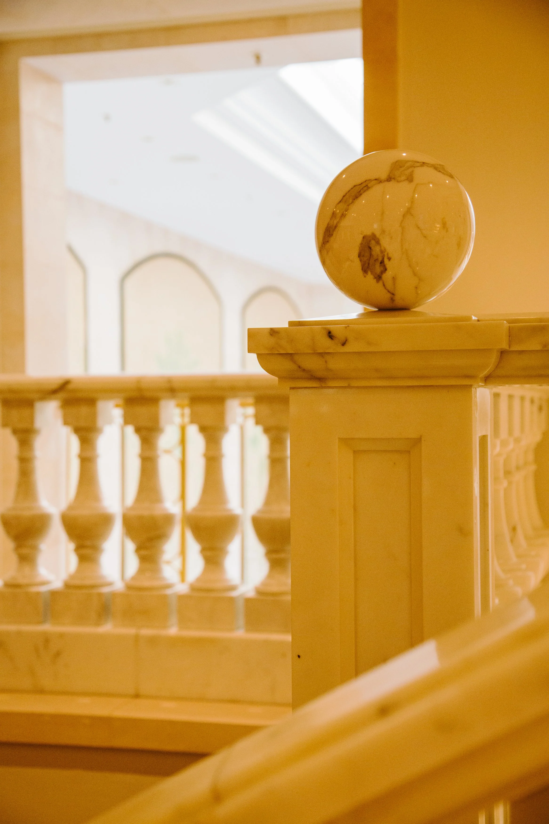 Close-up of a marble decorative sphere on a beige wooden railing, with a background of a staircase and balustrade. The scene is indoors with warm lighting.