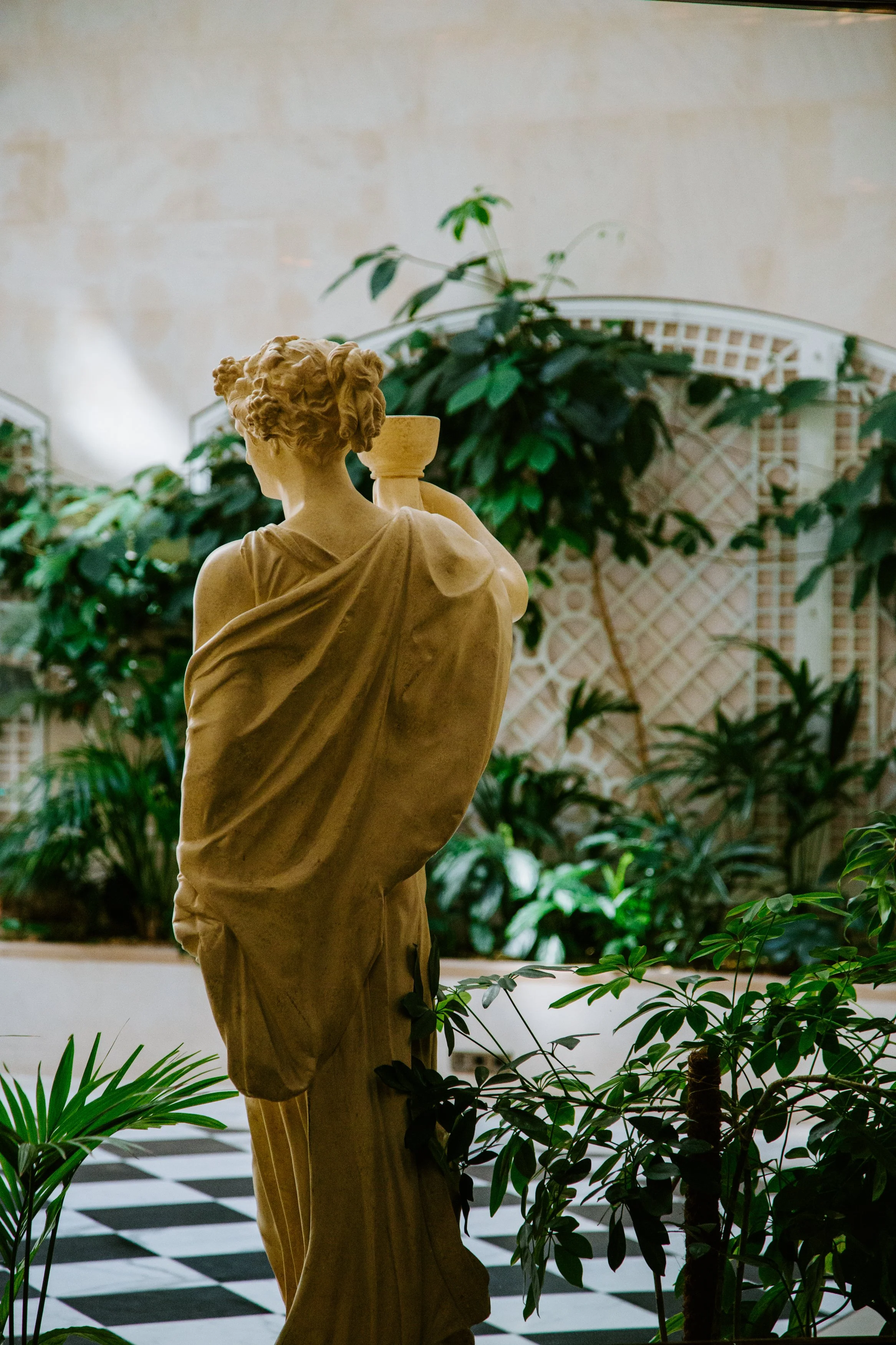 A classical marble statue of a woman viewed from behind, surrounded by green indoor plants and set against a background of a cream-colored wall with a lattice design and a potted plant. The floor features a black and white checkered pattern.