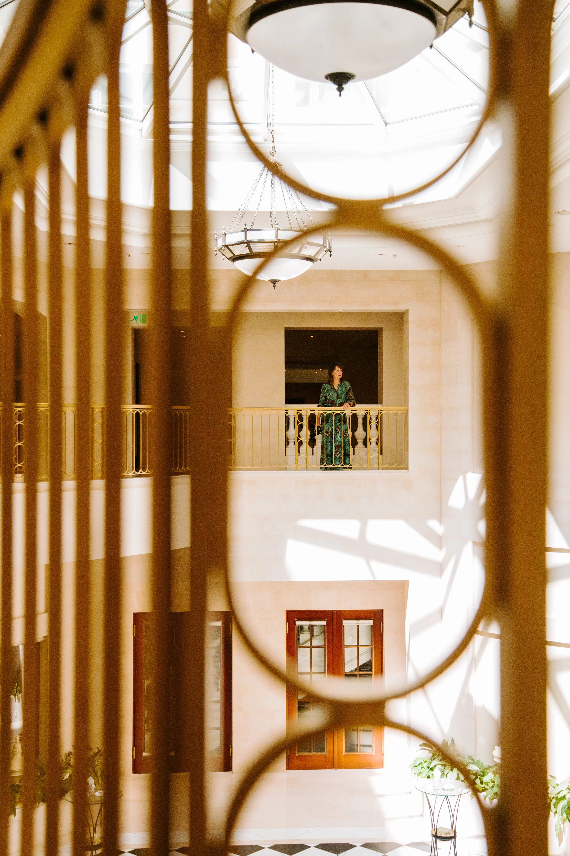 View through decorative iron railing of an interior space with a woman standing on a balcony, looking out. The space has a high ceiling with a large chandelier and skylights, and wooden doors on the wall below.