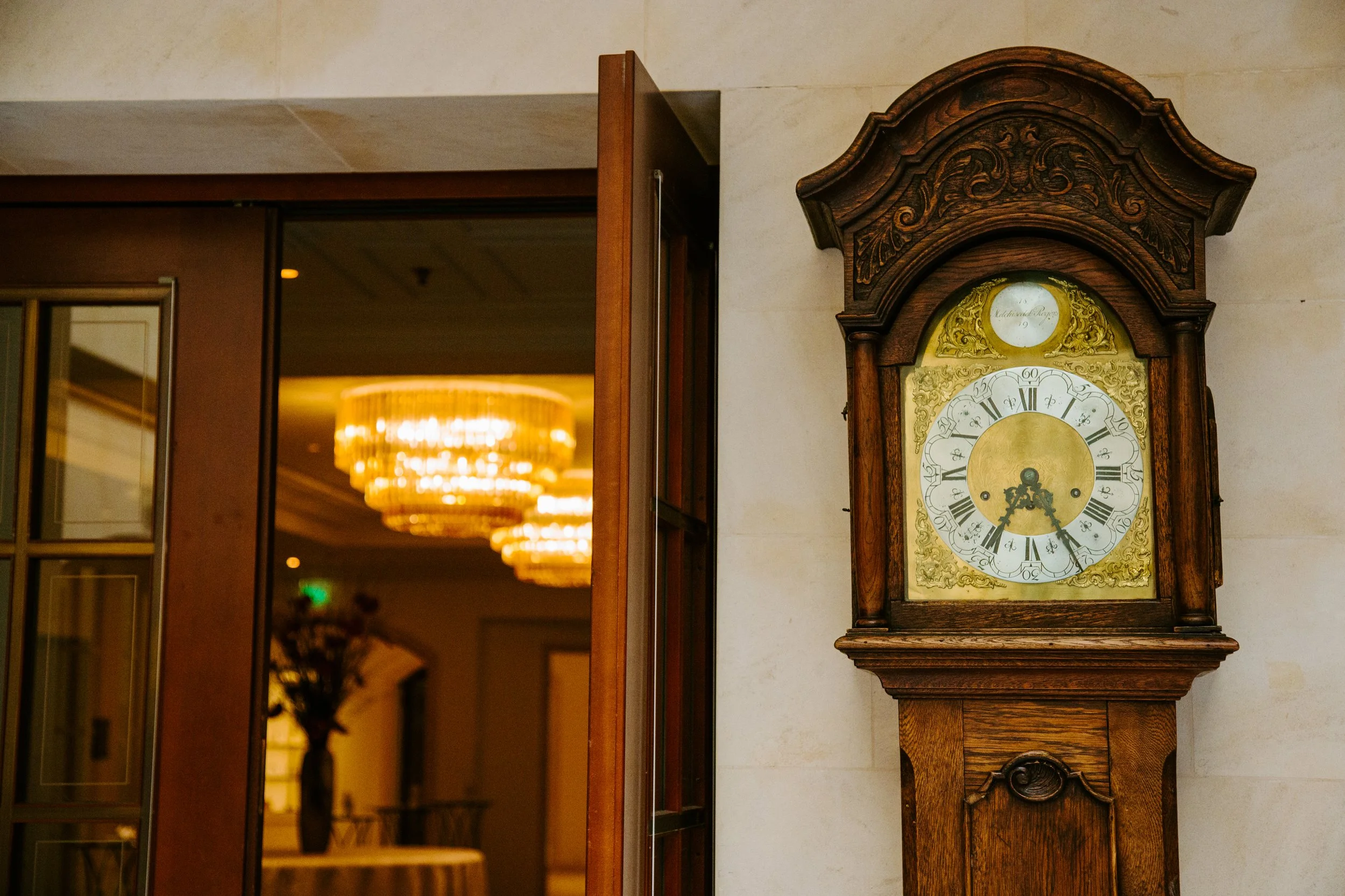 An antique wooden grandfather clock with a decorative face and Roman numerals, standing against a beige wall. In the background, a hallway with warm lighting and a chandelier is visible.