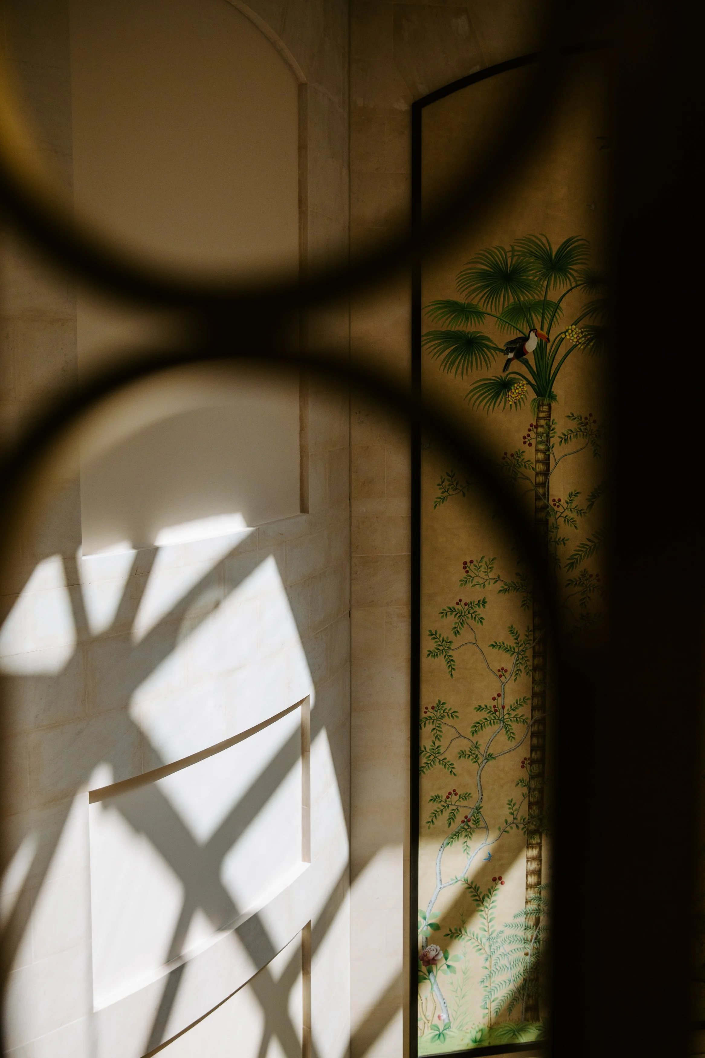 Interior view of a beige wall with a decorative panel featuring a painted tropical scene with lush green plants and a bird, partially obscured by a black circular object and shadows.