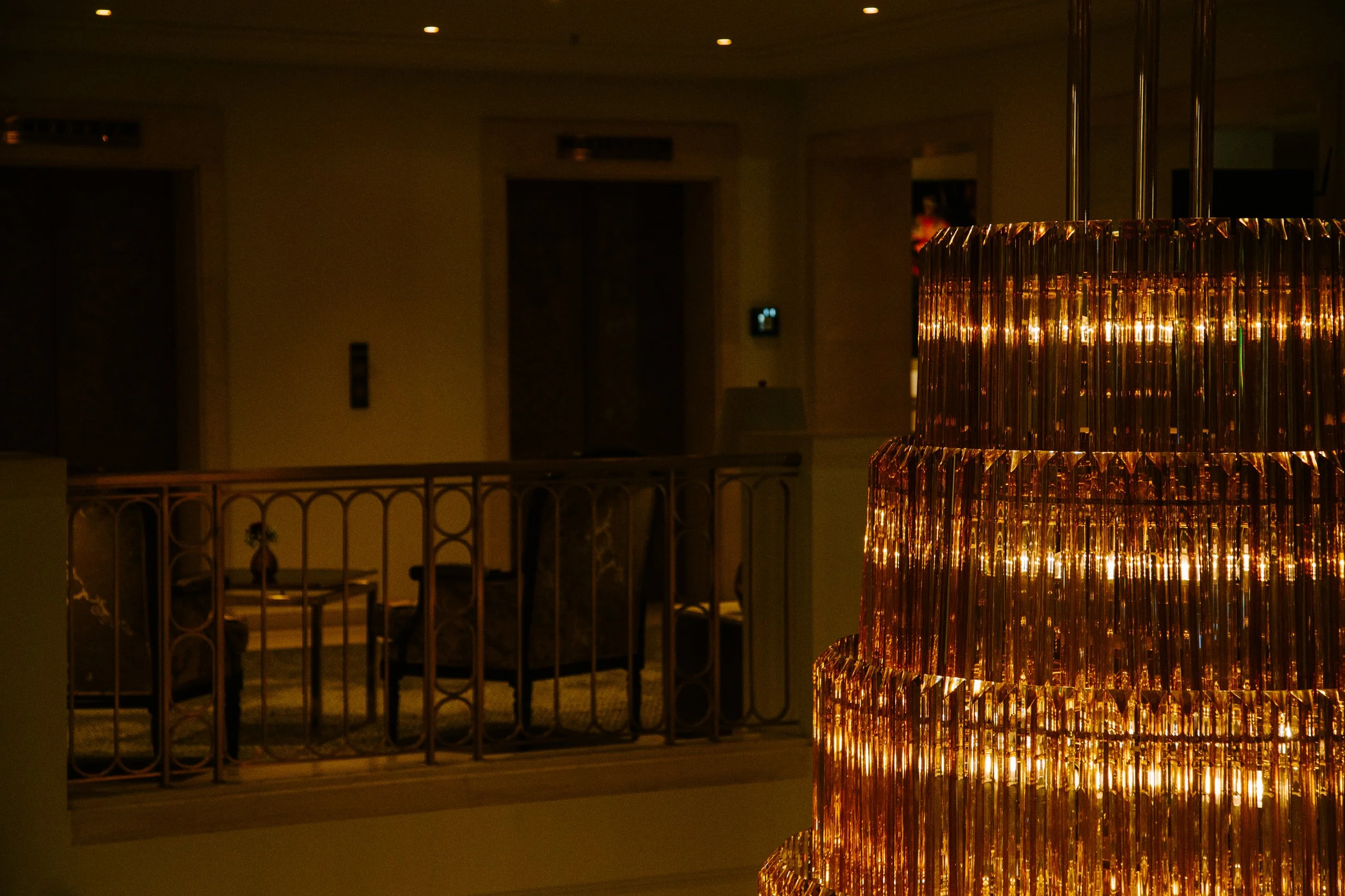 A dark indoor setting featuring a luxurious copper-colored chandelier with vertical glass rods. In the background, there's a staircase with a decorative railing and closed elevator doors, with dim lighting and a few recessed ceiling lights.