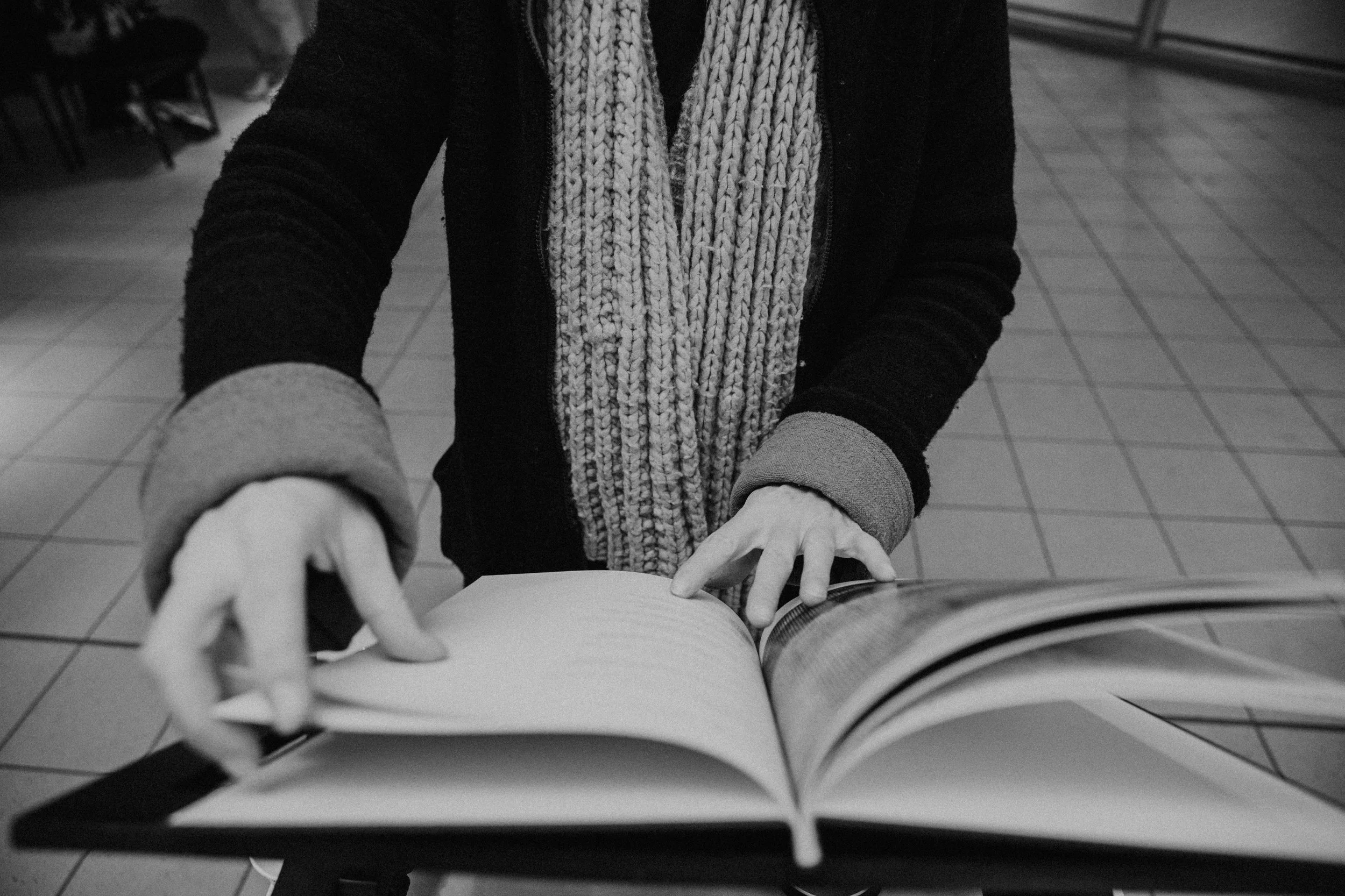 Black and white photograph of a woman in a striped scarf and dark clothing, leaning over and reaching towards an open book or magazine on a table. The setting appears to be indoors with tiled flooring and chairs in the background.