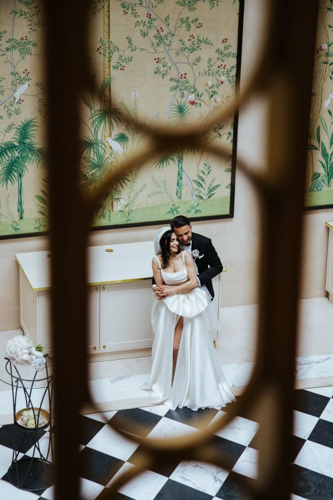 A bride and groom are smiling and embracing each other during their wedding, seen through a decorative iron fence with a floral and bird mural in the background.