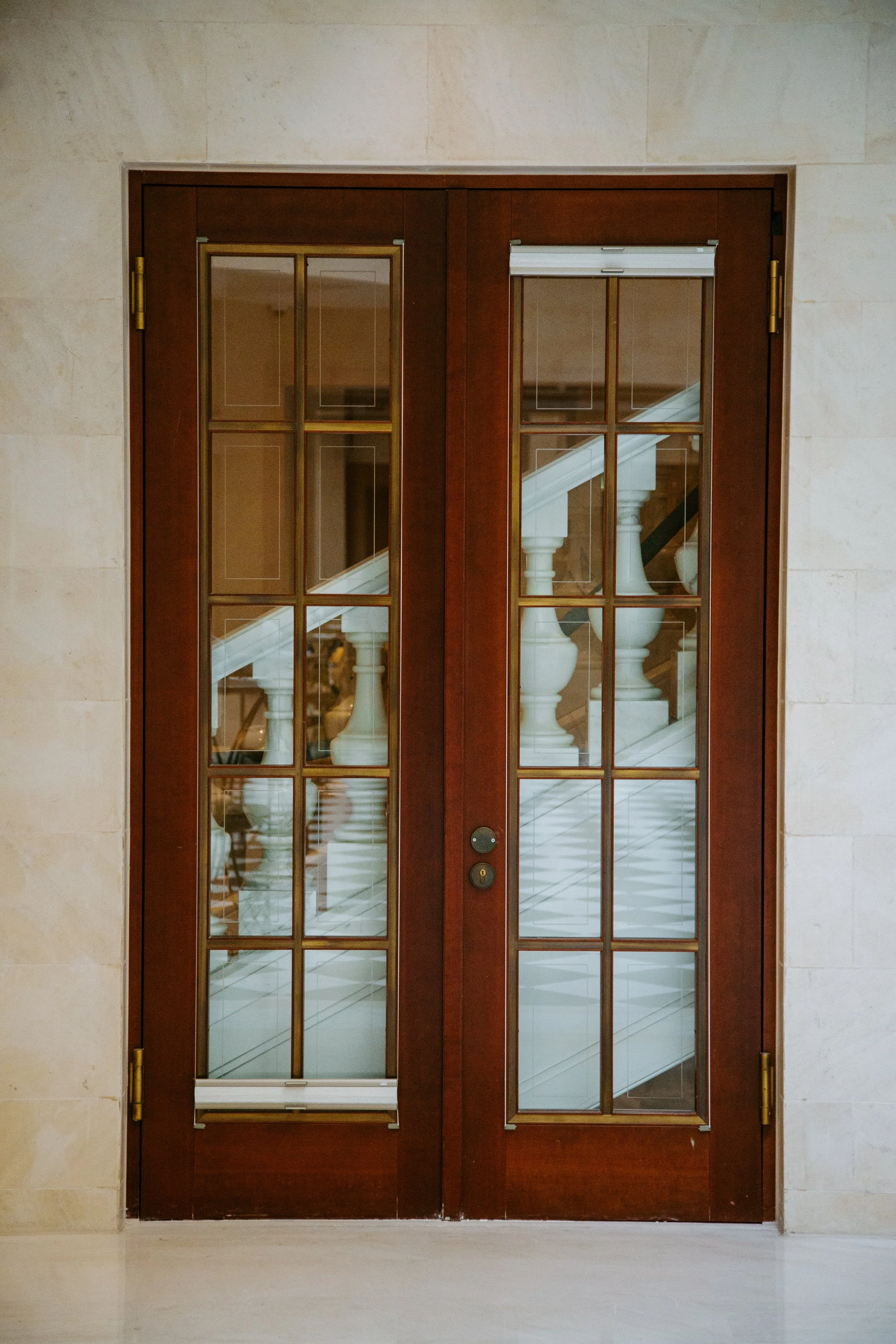 Wooden double glass doors with brass hinges and handles, leading into a building with a staircase visible through the glass panels.