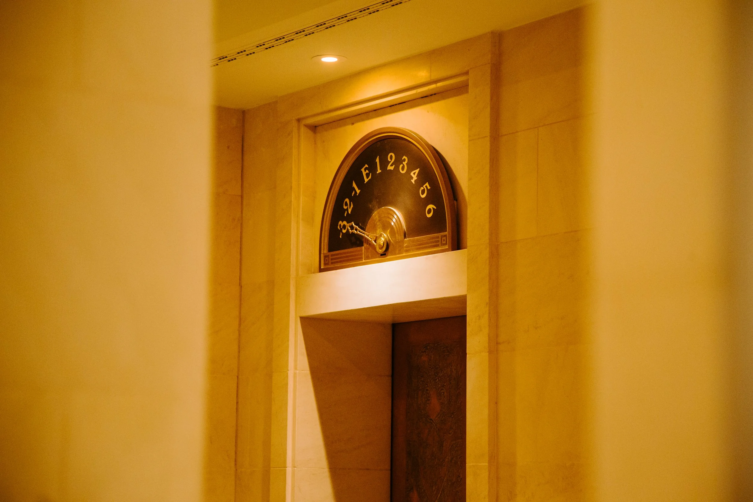 Close-up of a vintage clock with Roman numerals and ornate gold hands, mounted on a beige wall with warm lighting.