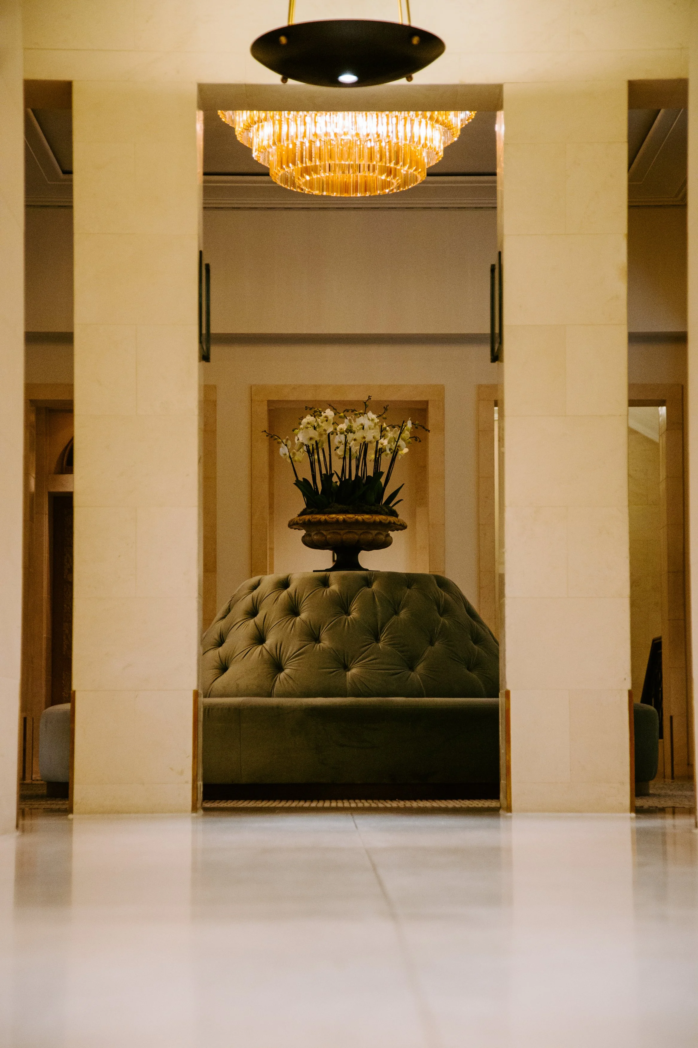 A chandelier hanging above a beige sofa with pillows. A large vase with white orchids on a table behind the sofa in an elegant interior.
