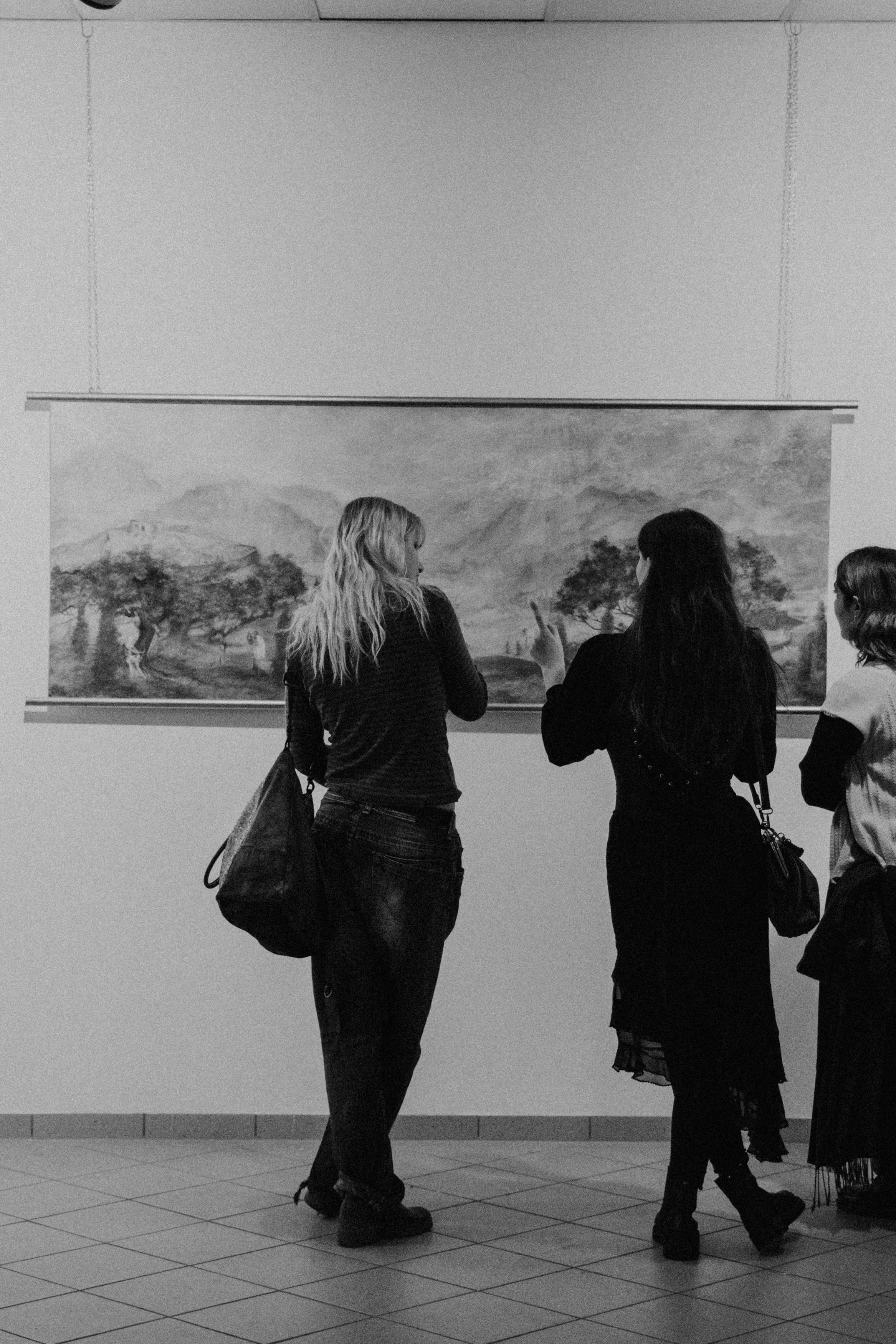 Three women standing in front of a landscape painting in a gallery, one of them is taking a photo with her phone.