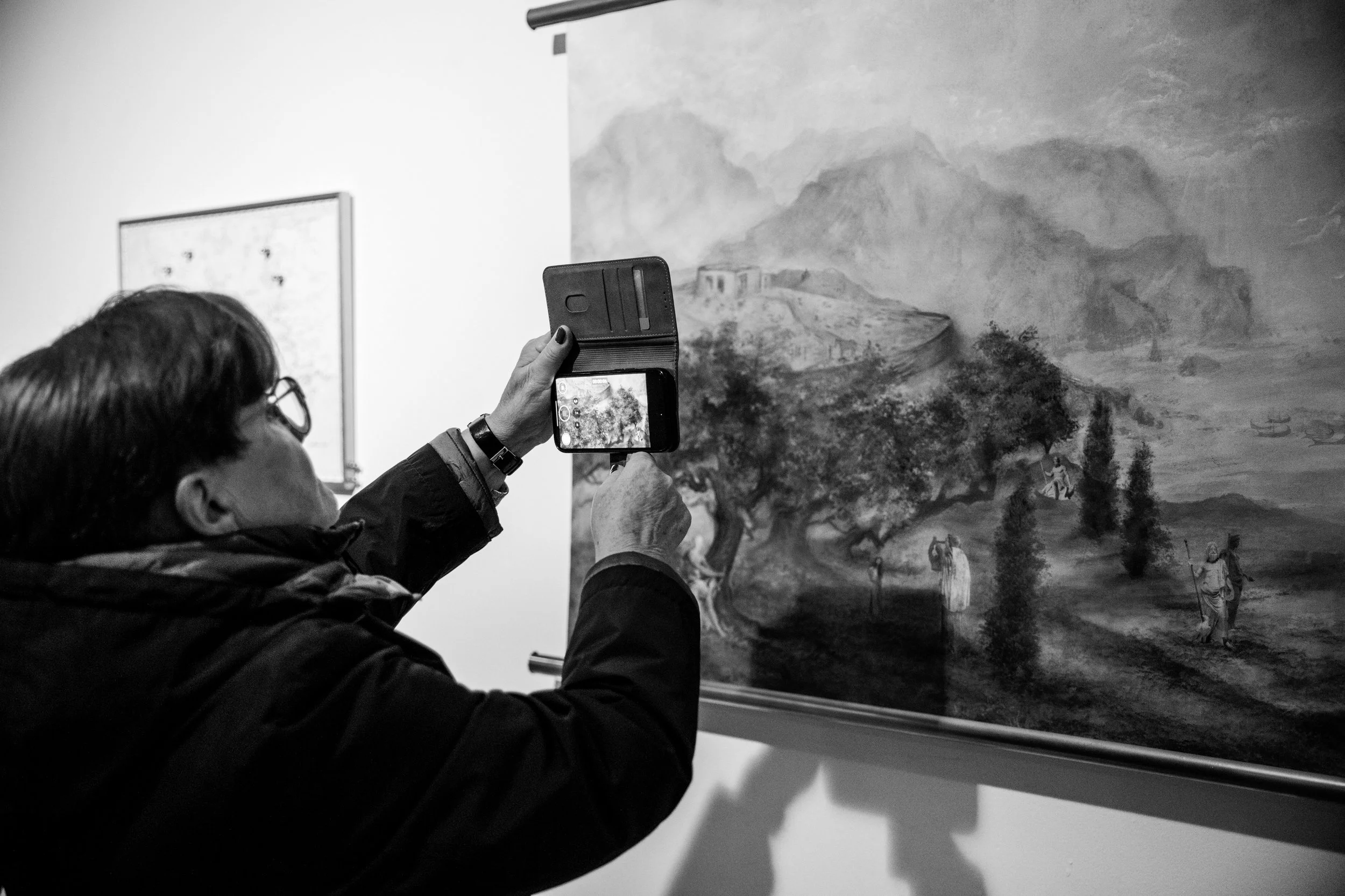 A person capturing a photo of a landscape painting with a smartphone in an art gallery.