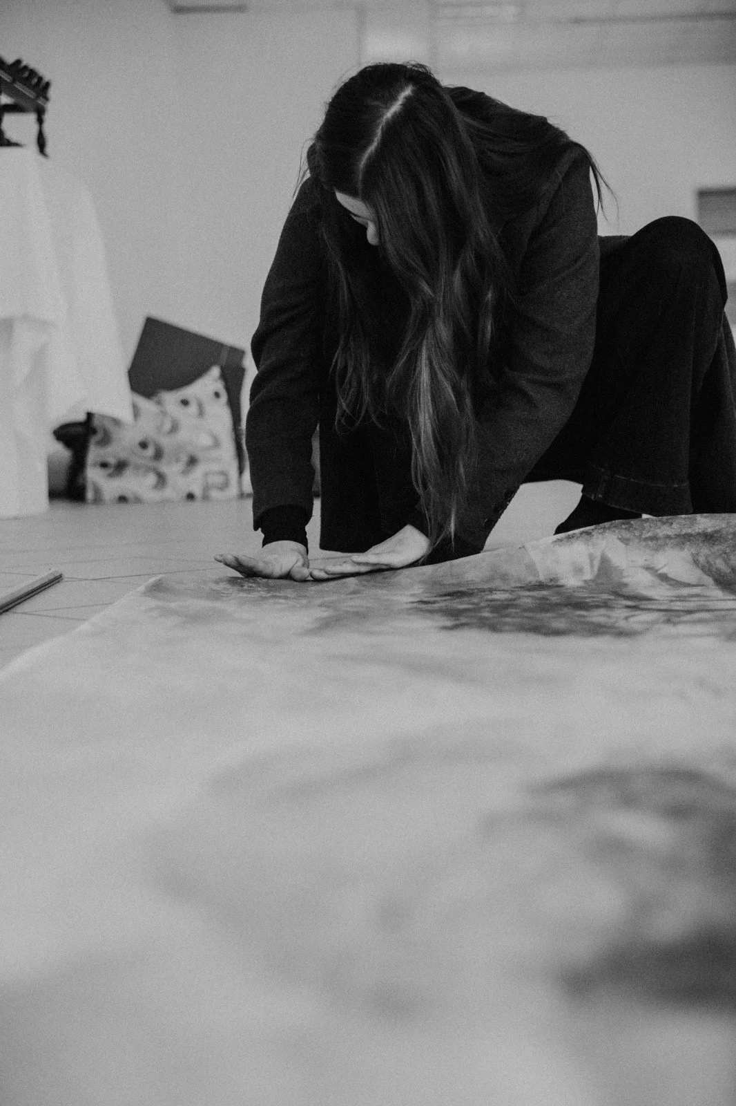 A woman with long dark hair bends over on her hands and knees, looking down at a large sheet or canvas on the floor in a room with art supplies and artwork in the background.