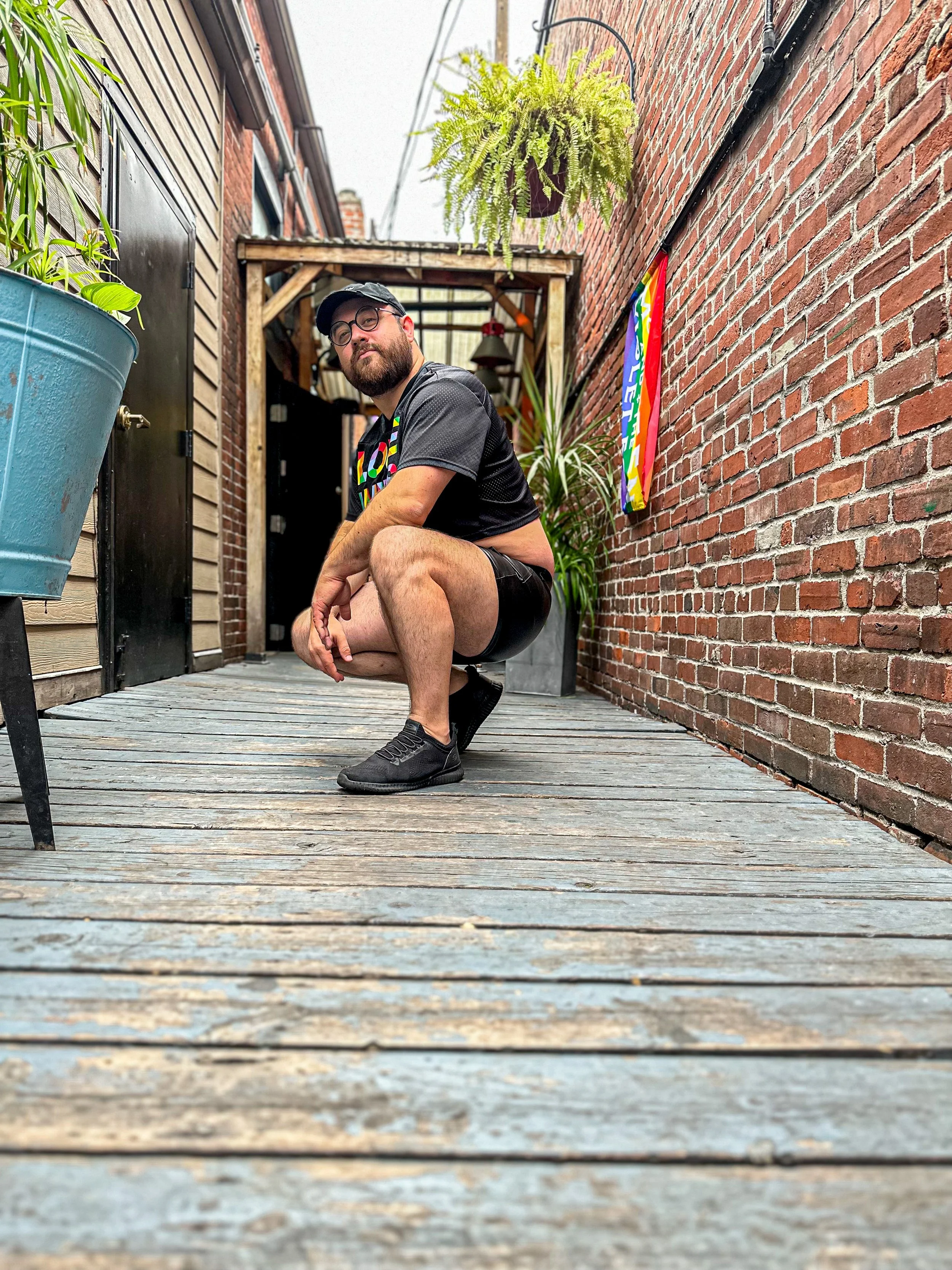 A man with a beard, glasses, a black cap, black t-shirt with colorful text, and shorts crouches on a wooden deck in an alleyway between brick buildings, with plants and a flag in the background.