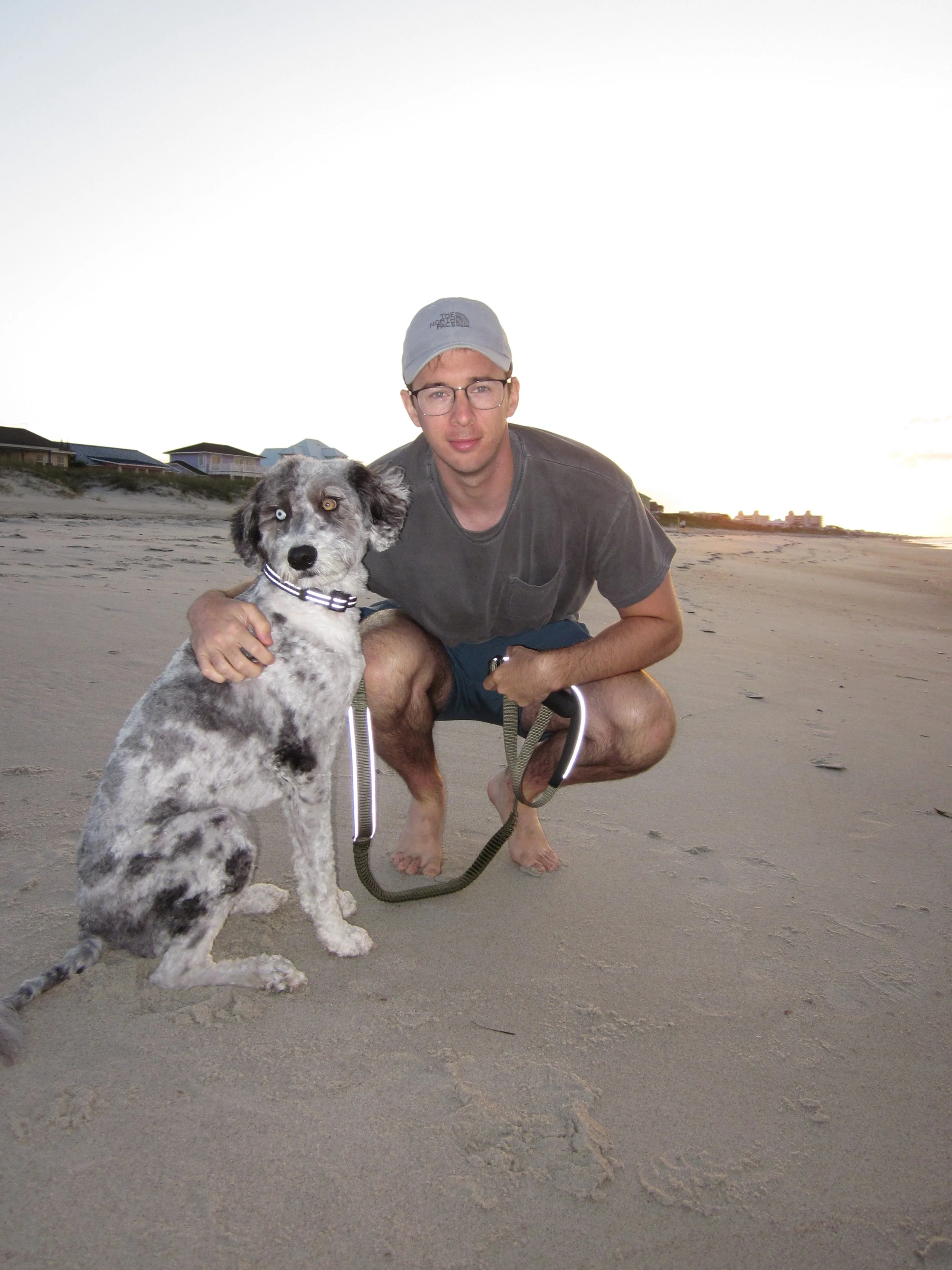 A young man with glasses, wearing a gray baseball cap and a dark T-shirt, crouches on a sandy beach at sunset, holding a black and white Border Collie x Poodle puppy with one blue eye and one brown eye on a leash.