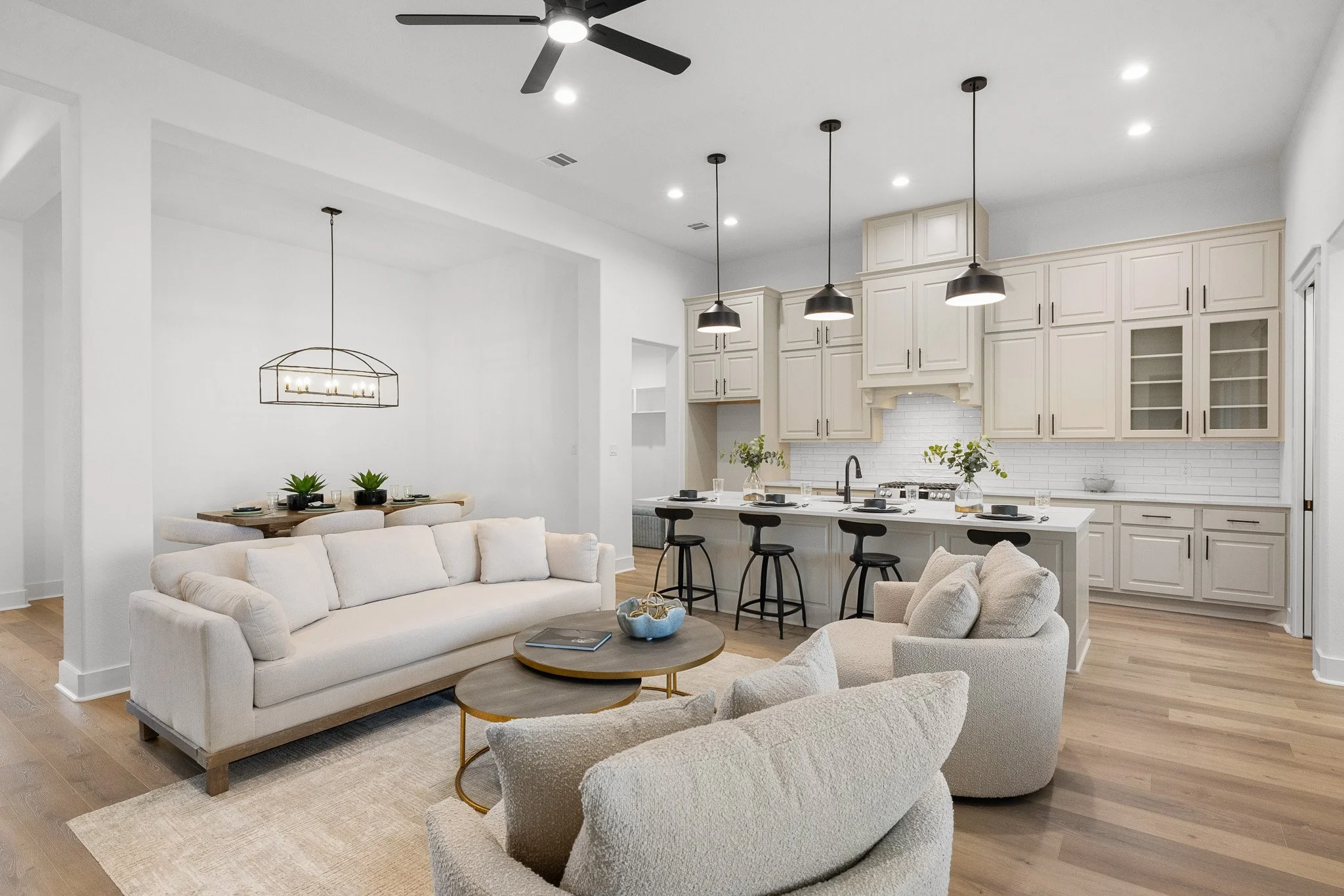 Open-concept living room and kitchen with white walls, beige and cream furniture, black pendant lights, and a ceiling fan.