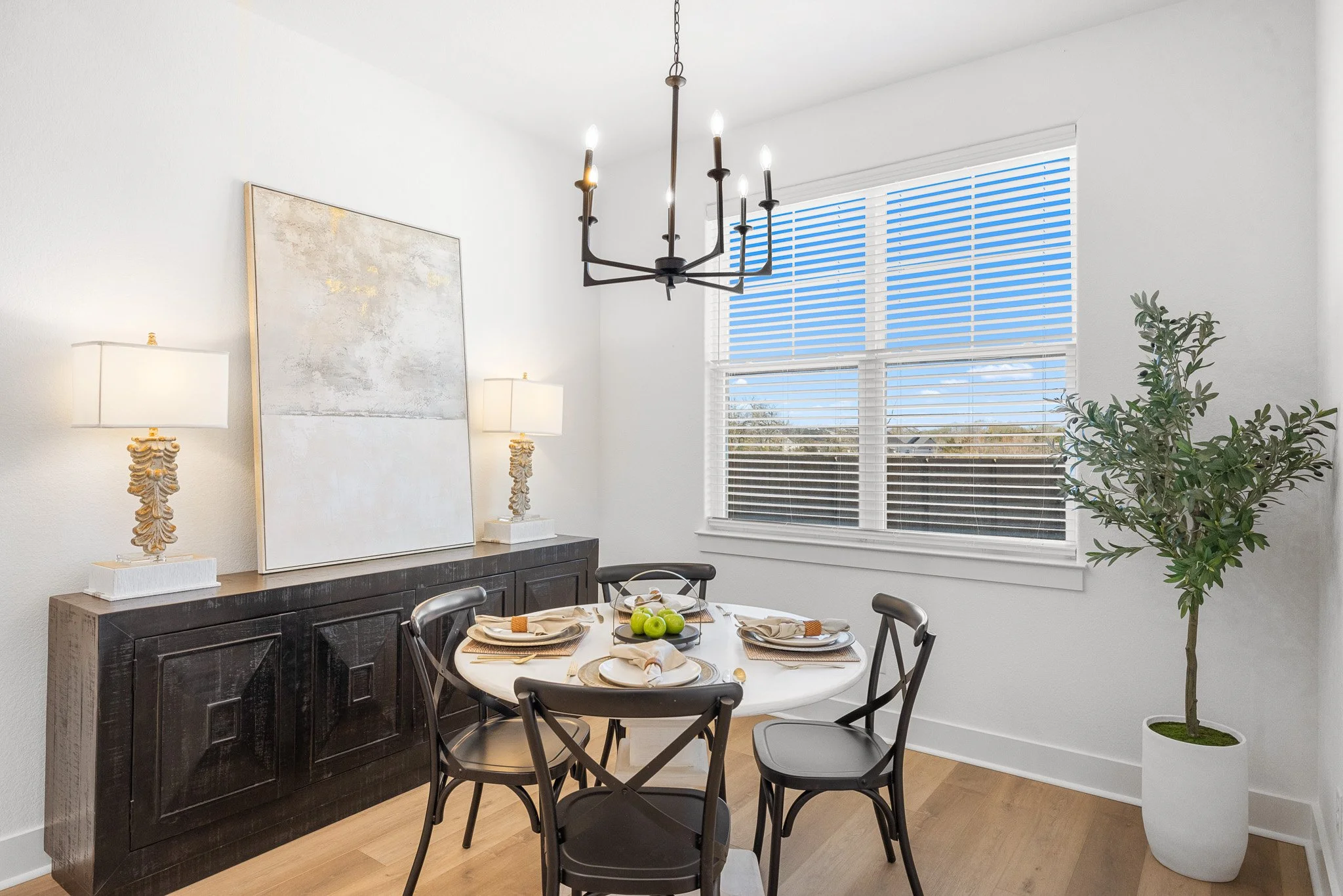 A dining room with a round table set with four place settings, a black buffet with decorative lamps and artwork, a large window with blinds, a chandelier, and a tall potted plant.