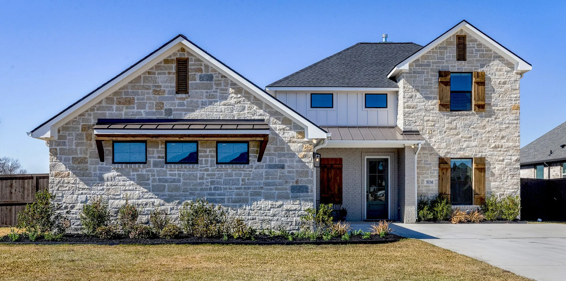 Front view of a modern house with stone and siding exterior, multiple windows with wooden shutters, a small front yard with bushes, a concrete driveway, and a blue sky.