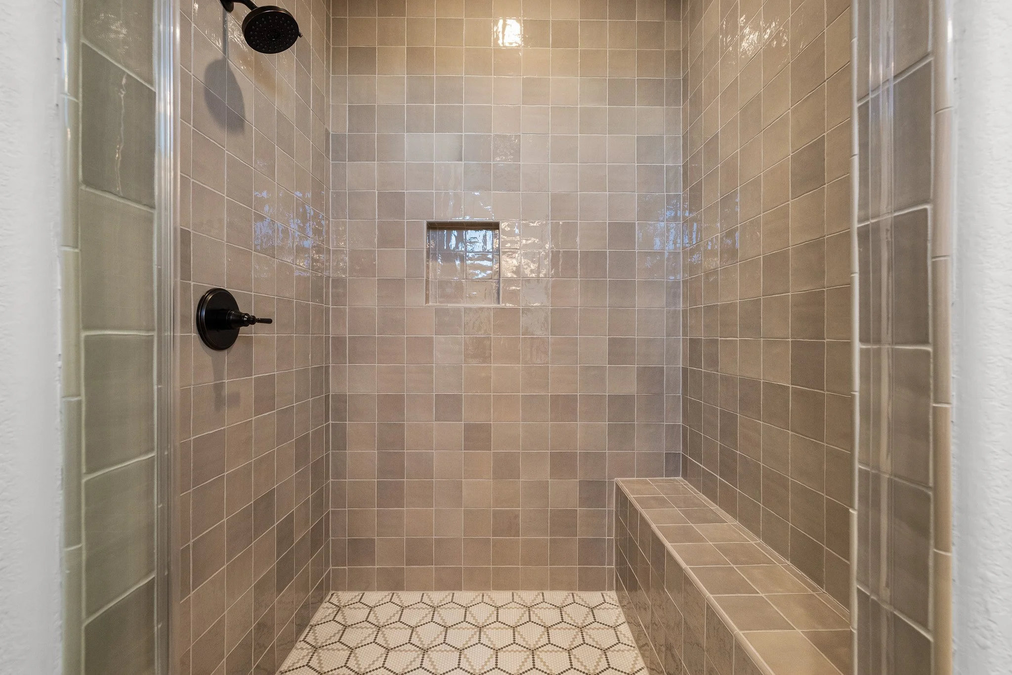 Inside a tiled walk-in shower with a black showerhead and control valve, beige wall tiles, and built-in corner seating.