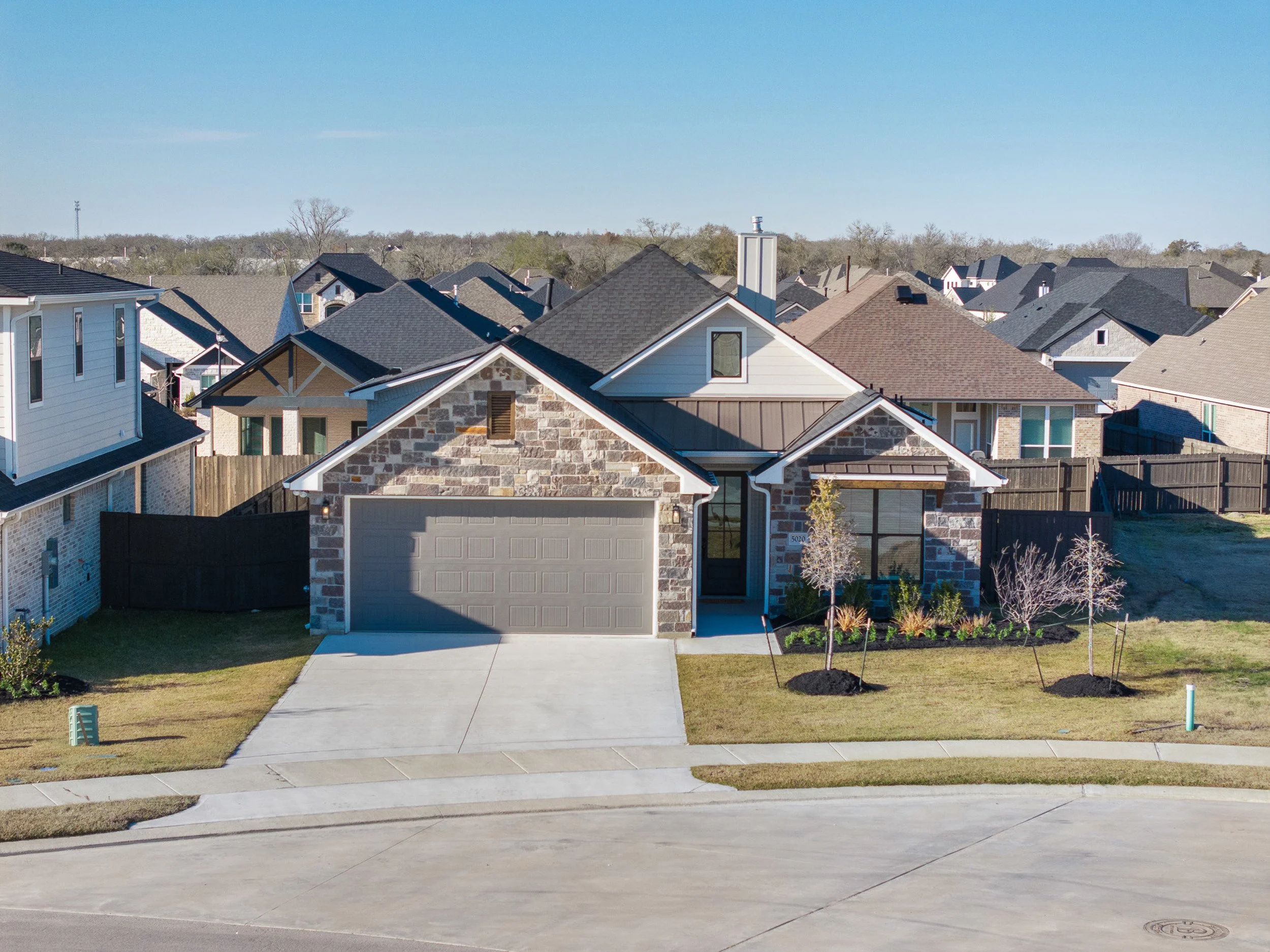 Front view of a modern house with a stone and siding exterior, a two-car garage, a front lawn with young trees, and neighboring houses in the background under a clear blue sky.