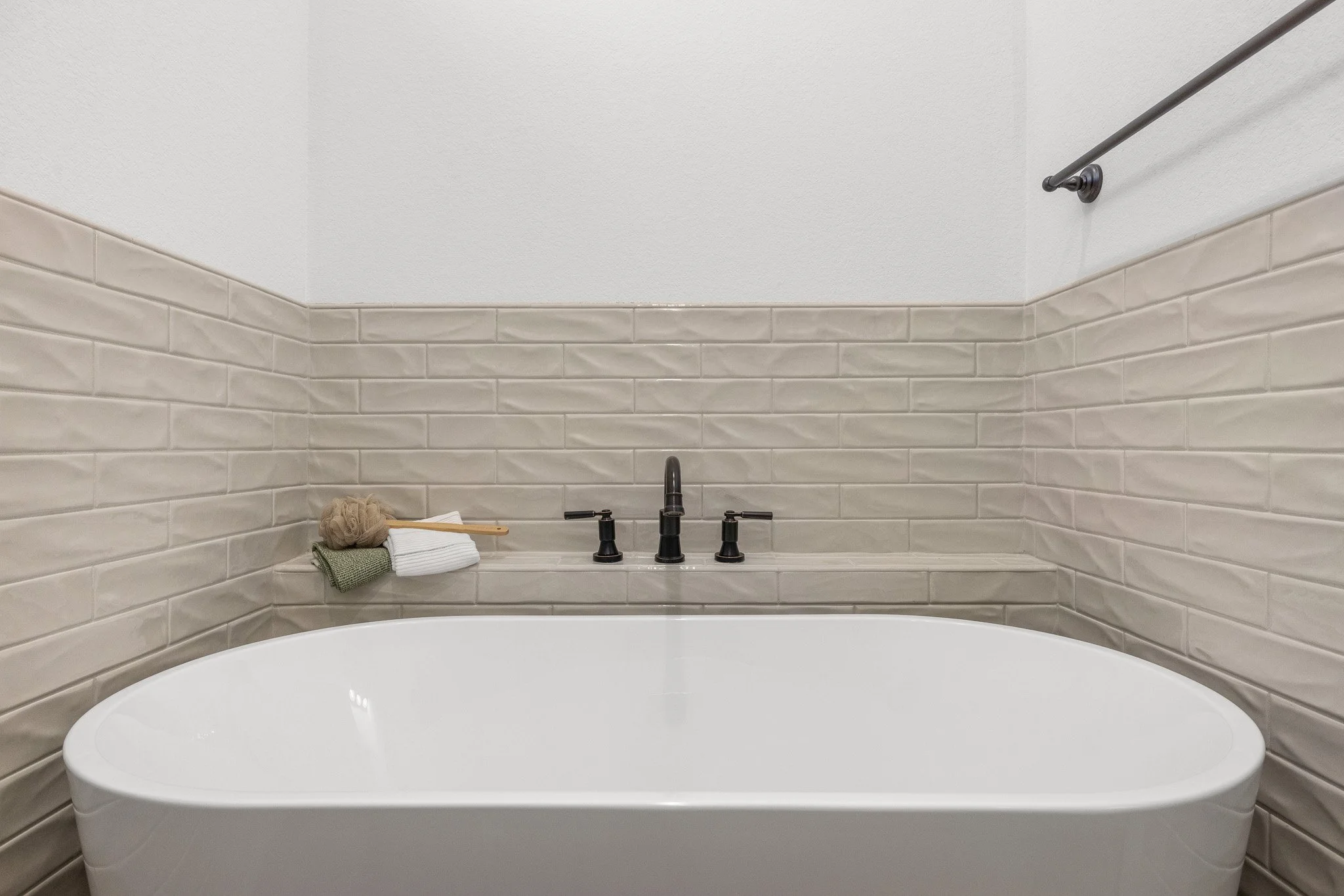 A bathroom with a bathtub in the foreground and beige tiled walls behind it. There is a black faucet and three handles for the bathtub. On the ledge, there are neatly folded towels and a bath sponge. A towel bar is mounted on the wall above the tiles