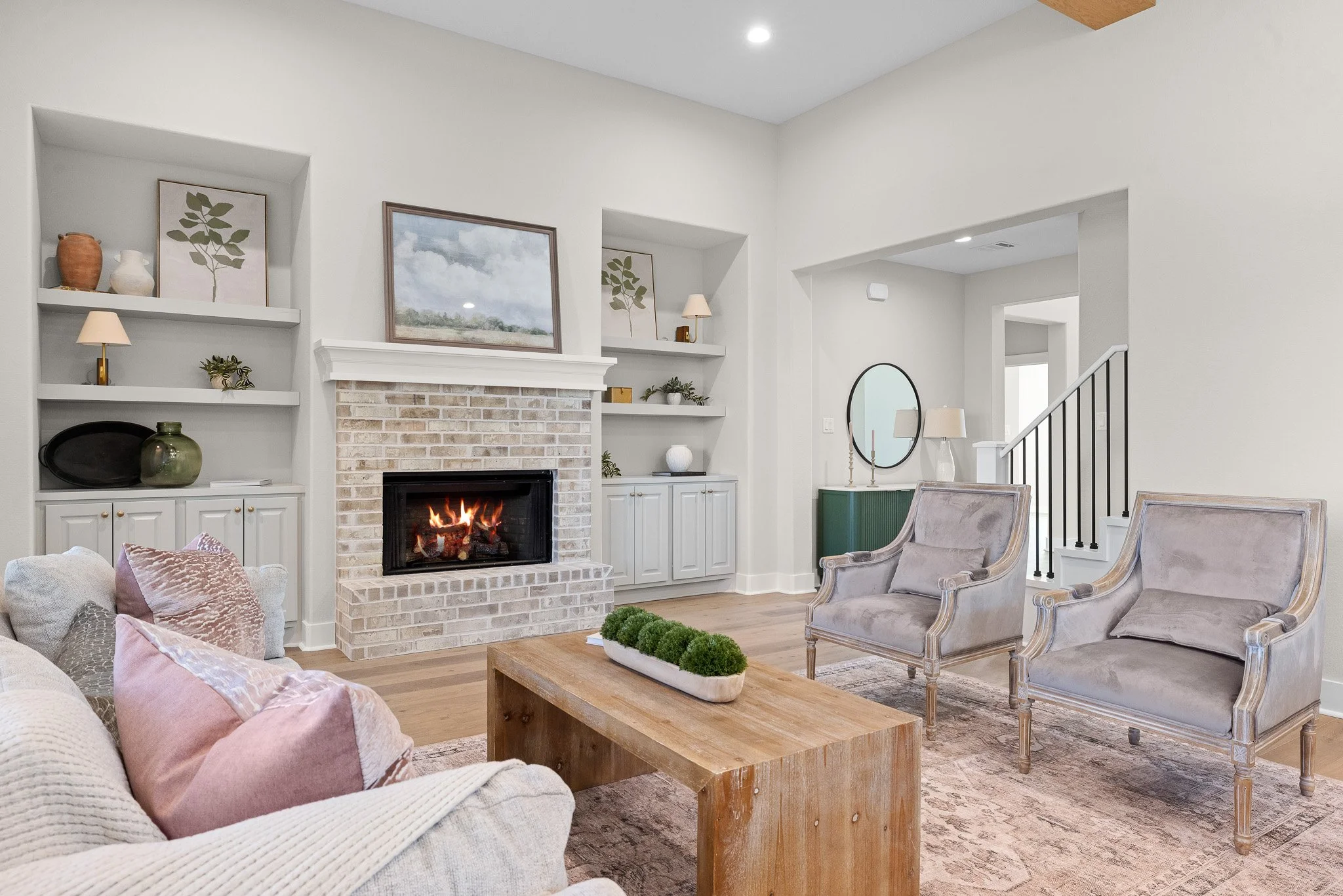 Living room with a brick fireplace, two built-in shelves, and artwork. There are armchairs, a wooden coffee table, and decorative items including lamps, vases, and plants.