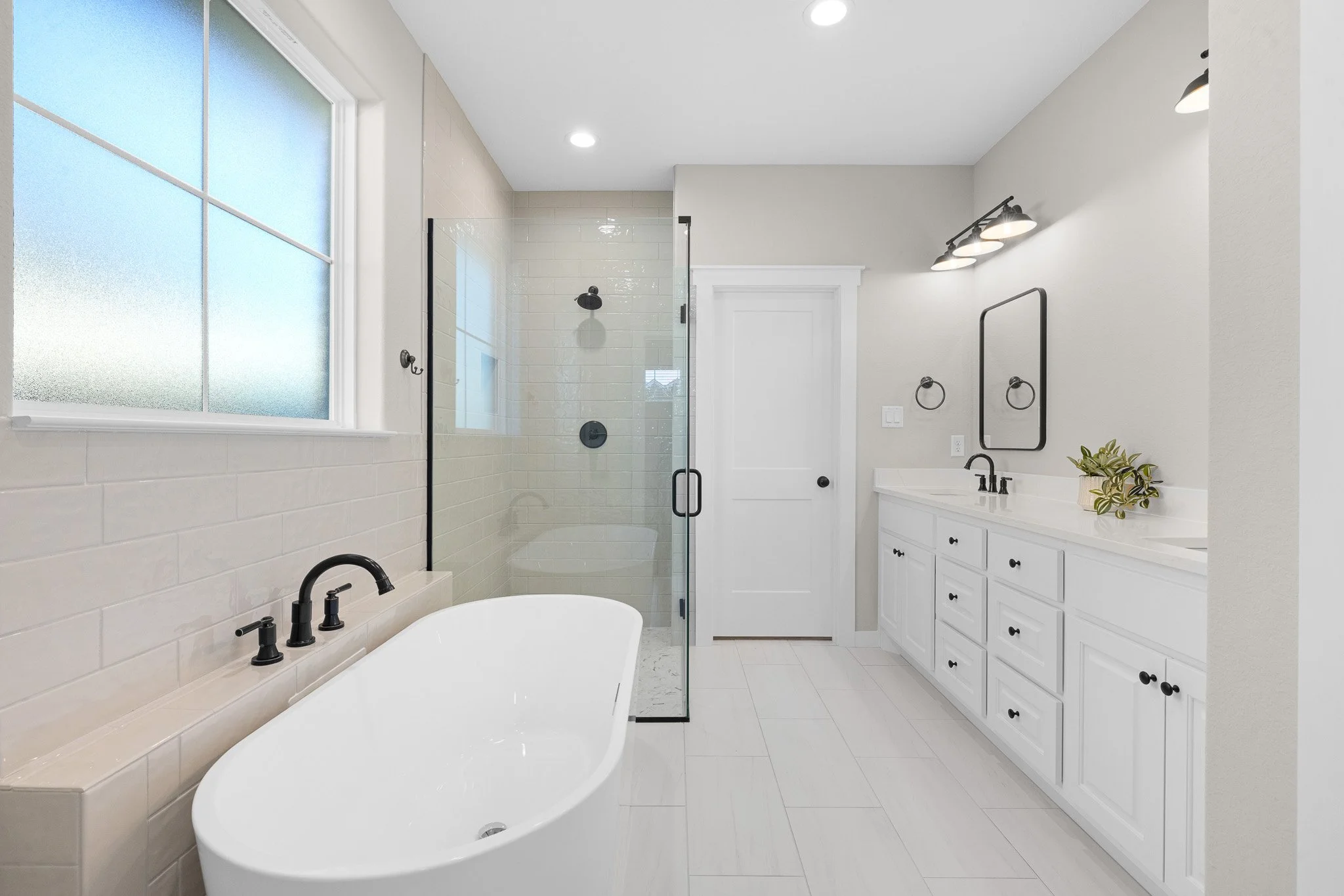 A modern bathroom with a white bathtub, black fixtures, a large frosted window, a glass-enclosed shower, white cabinets with black hardware, a black-framed mirror, and a small plant on the counter.
