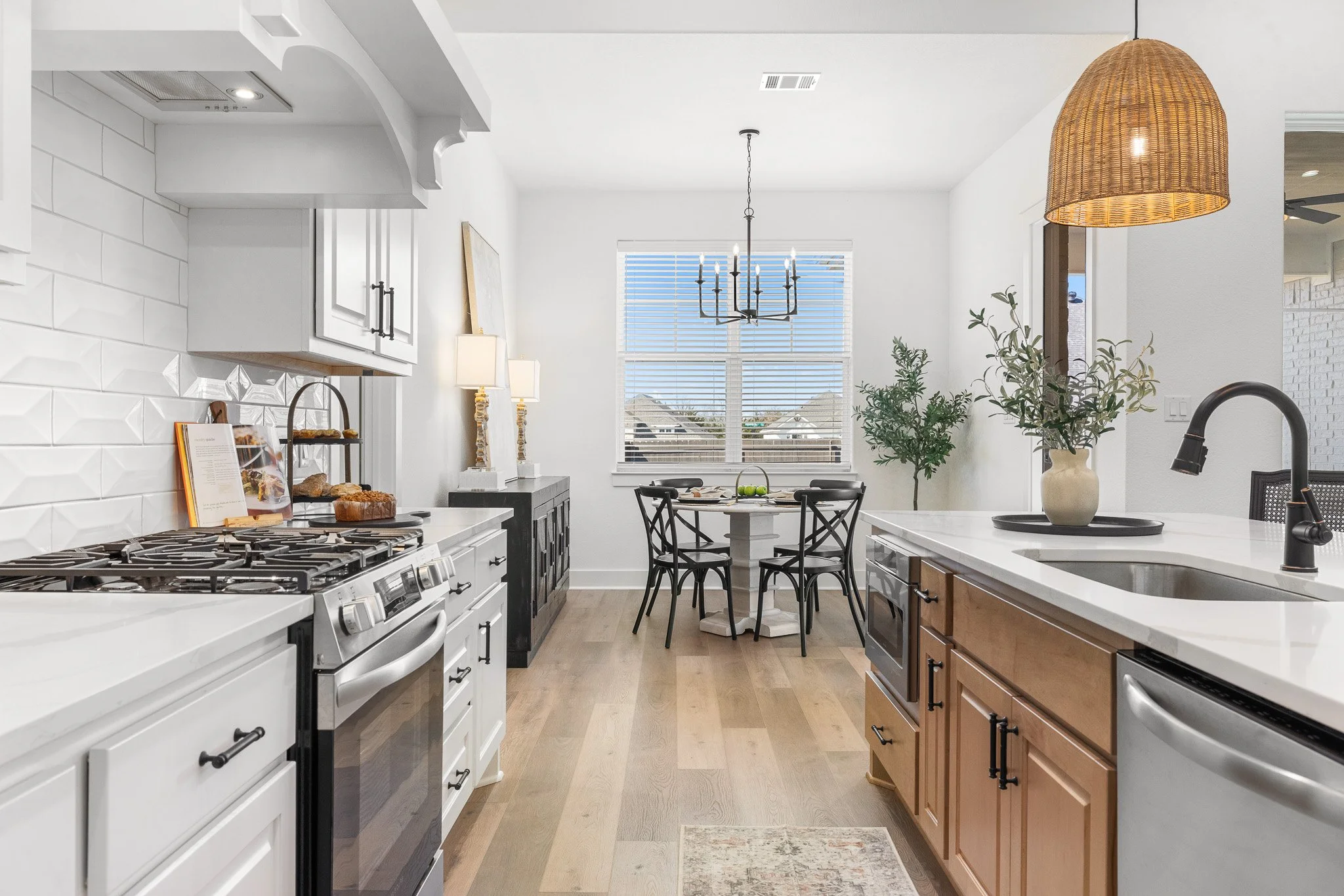 Bright modern kitchen with white and wood cabinets, stainless steel appliances, a dining table with four chairs, a large window with blinds, and various decorative items.