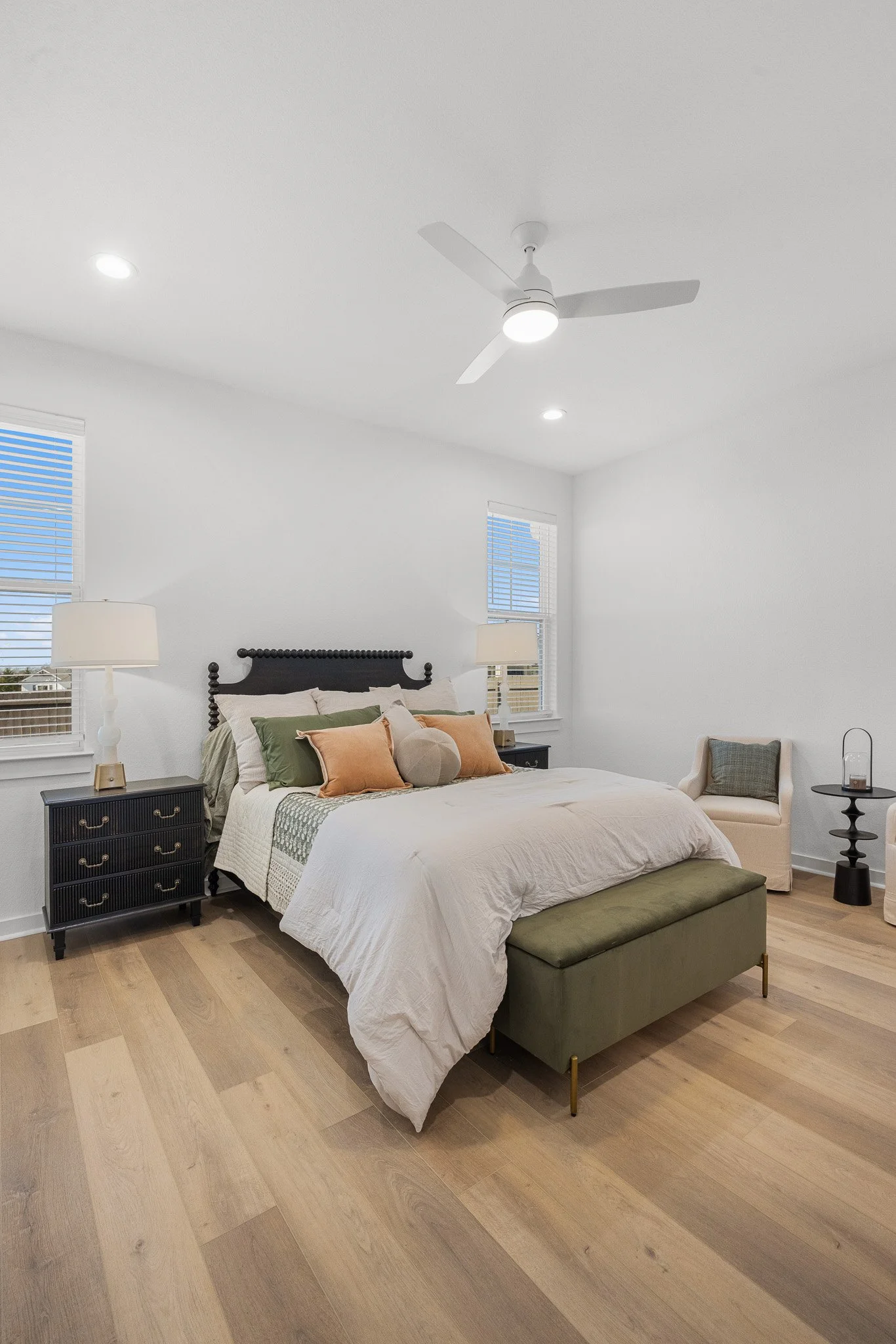 A bedroom with a large bed with beige bedding, multiple pillows, and a dark headboard. There are two black nightstands with lamps on either side of the bed. A light green bench is at the foot of the bed. The room has two windows with white blinds, an