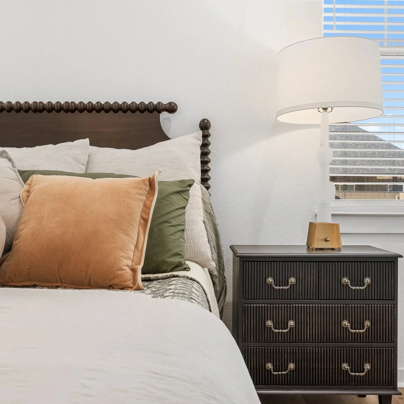 A bedroom with a dark wooden headboard, beige and green pillows on a white bedspread, a black nightstand with a vintage brass handle, a white table lamp with a cylindrical shade, and a window with blinds showing outdoor houses.