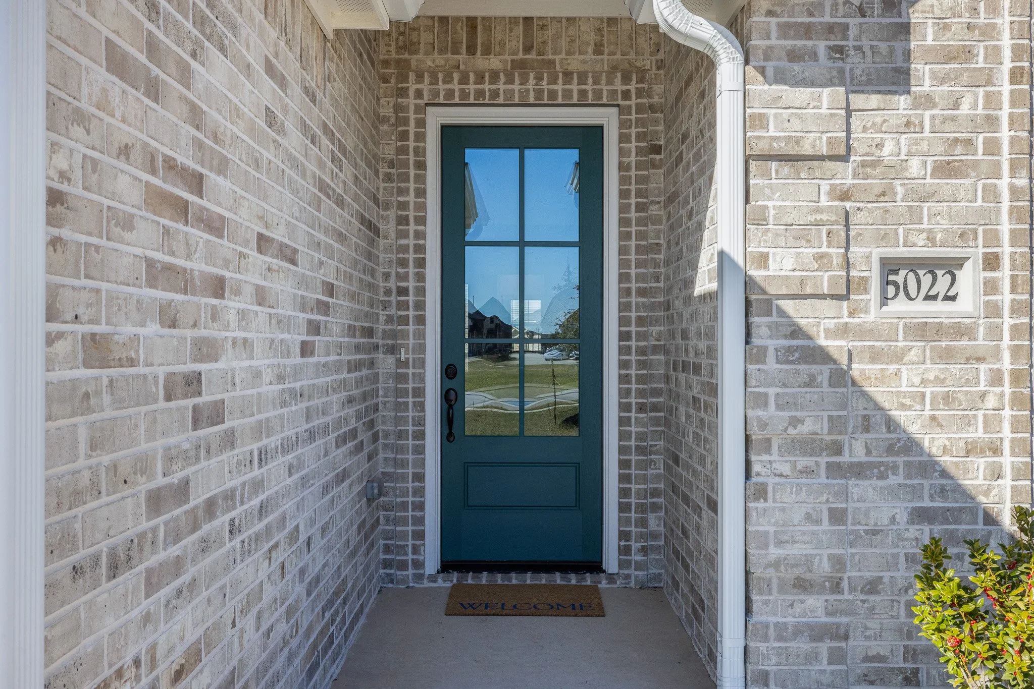 Front porch of a house with a brick wall, a teal front door with glass panels, a welcome mat, and some greenery on the right.