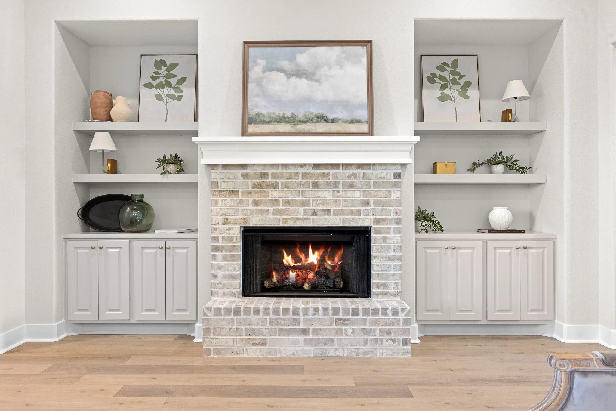 Living room with a brick fireplace with a fire, white built-in shelves on each side decorated with vases, plants, and lamps, and framed artwork above the mantel. Light wood floor and white walls.