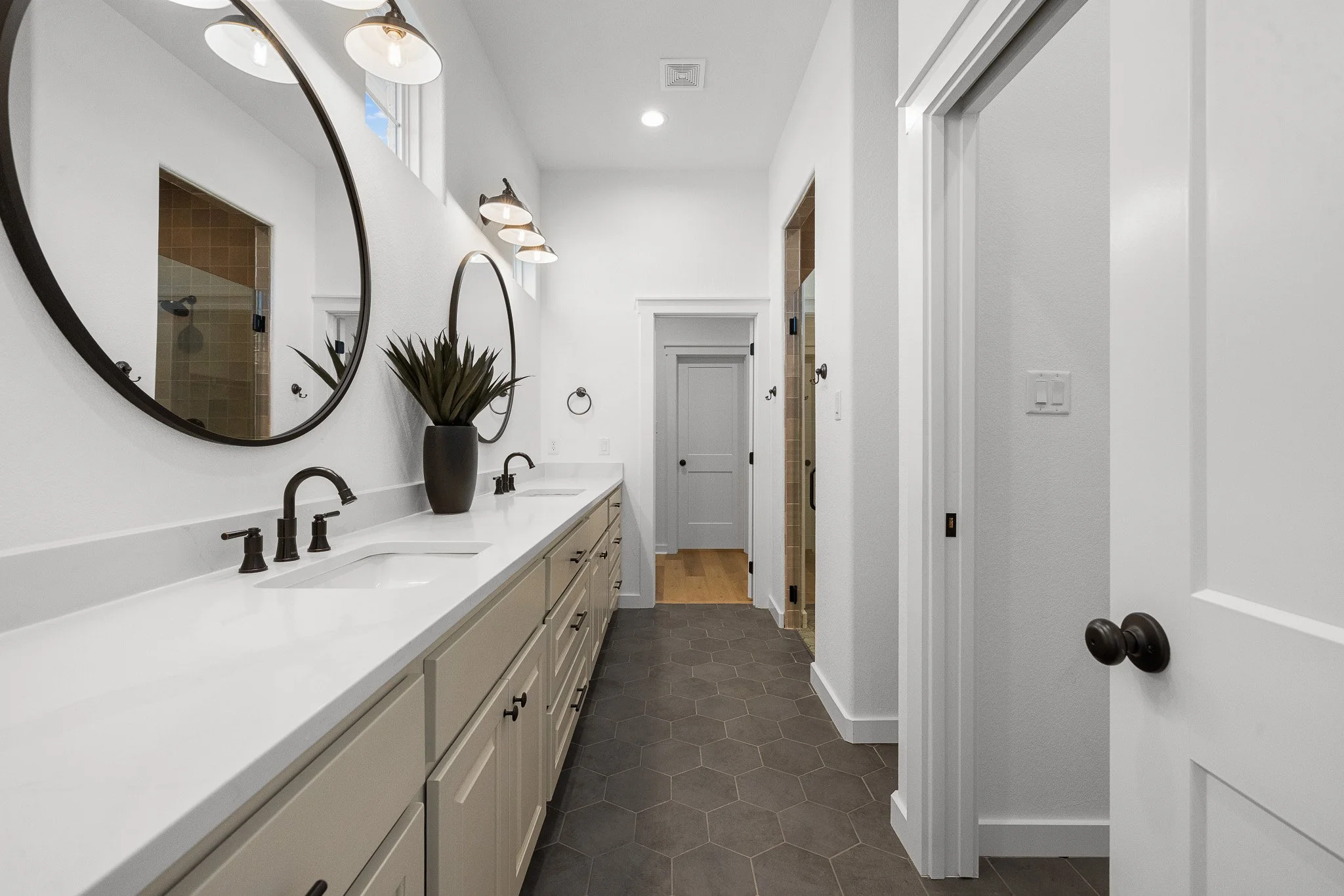 Bathroom with white cabinets, black fixtures, two round mirrors, potted plant, hexagon dark gray tiles, and multiple light fixtures on a white wall.