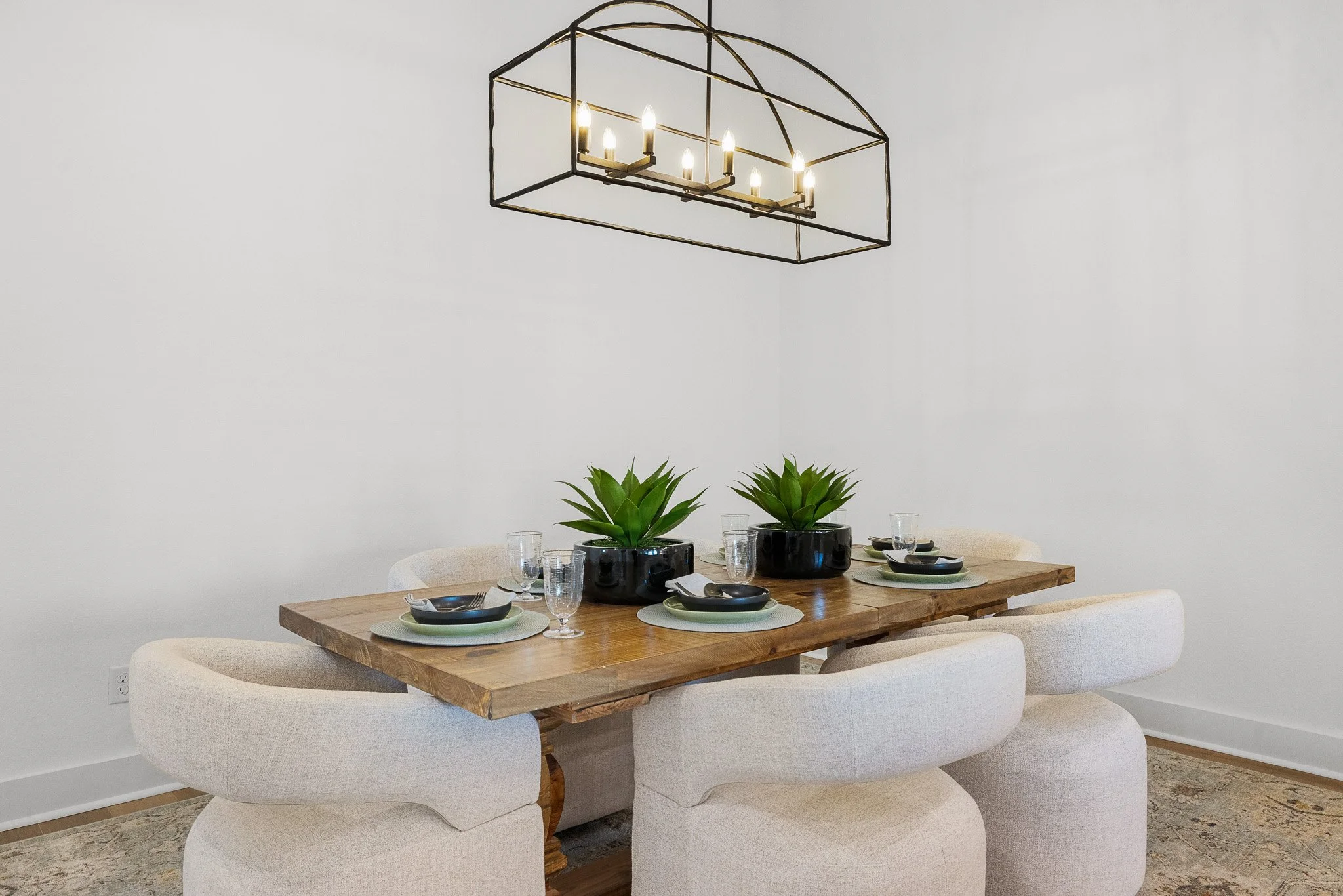 A dining room table set for four with black plates, green placemats, and drinking glasses, decorated with two potted green plants, and lit by a black metal chandelier with multiple candle-style lights.