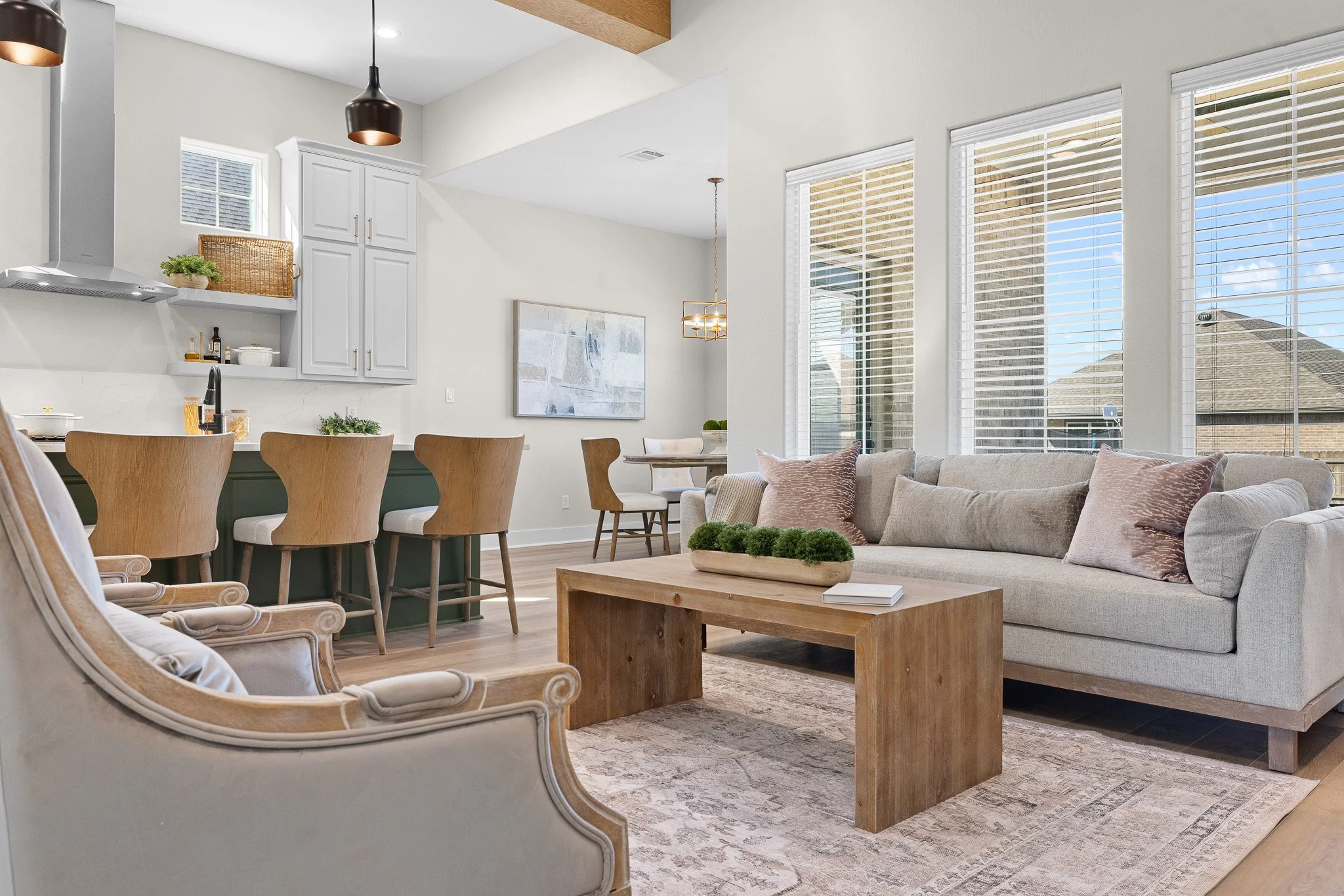 Open-concept living room and kitchen with large windows, beige sofa, wooden coffee table, armchair, and dining area with four wooden chairs.