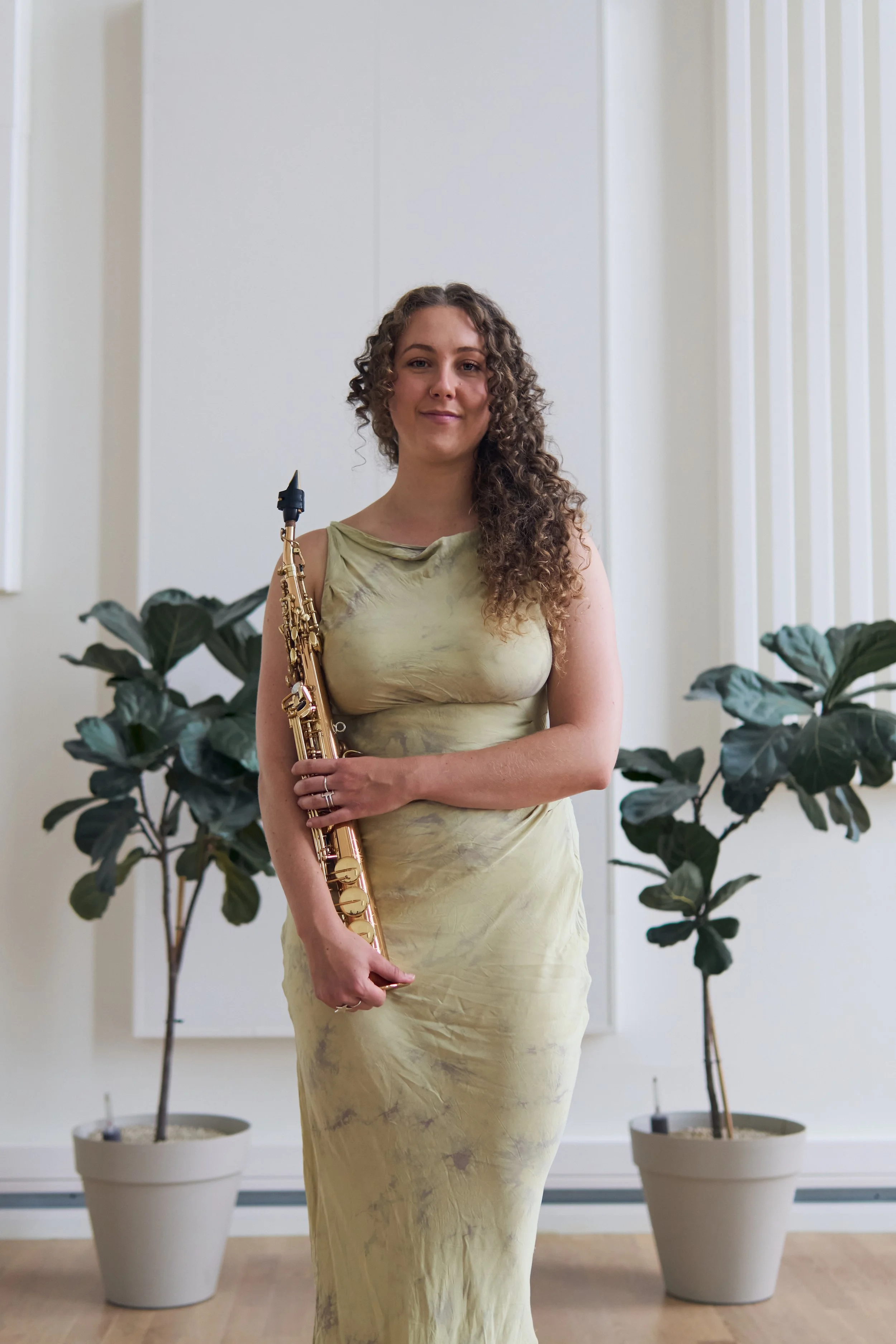 A woman with curly hair wearing a light yellow dress, holding a gold-colored alto saxophone, standing in front of potted plants and a white wall with vertical panels.