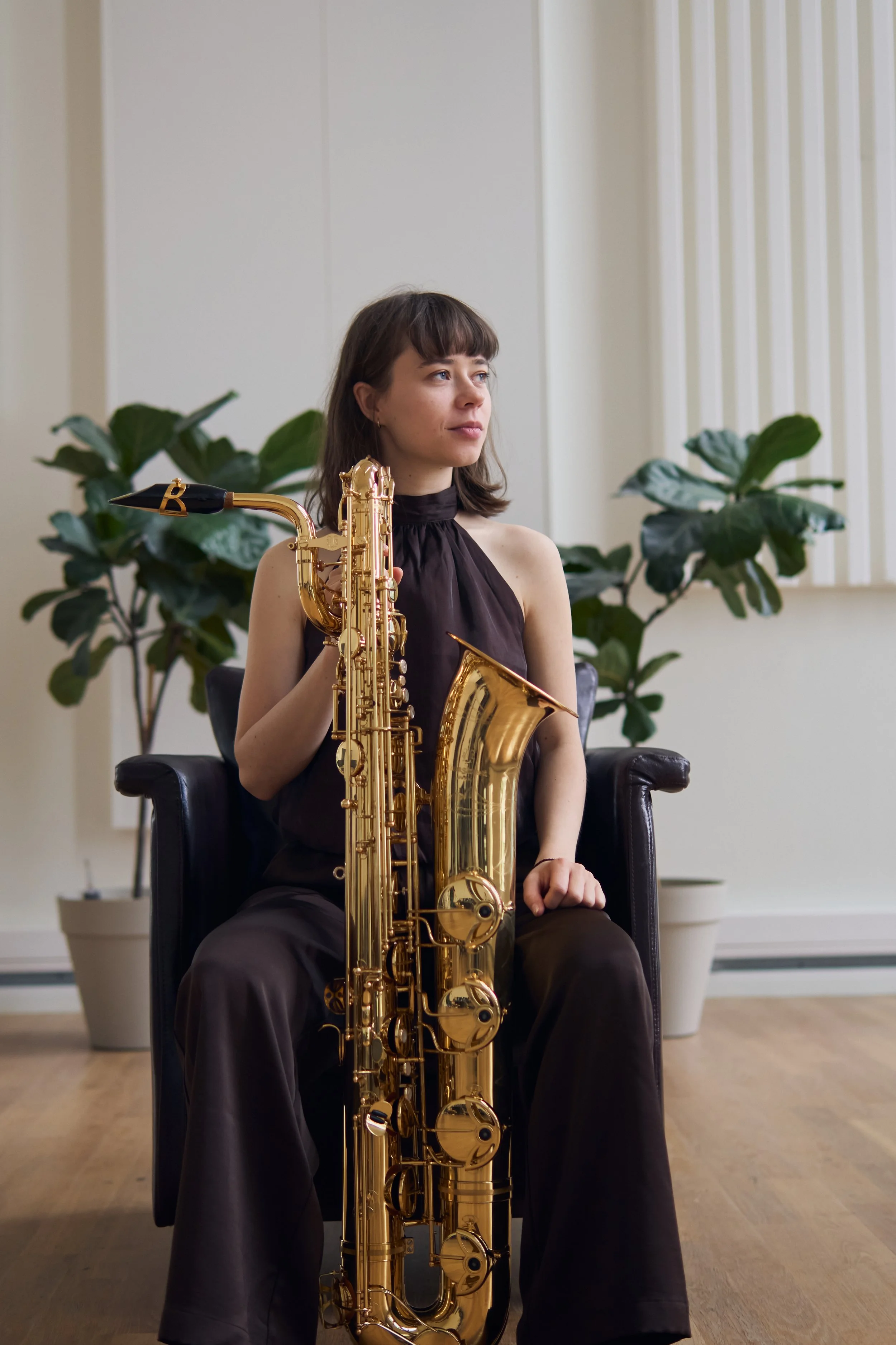 A woman sitting in a black leather armchair holding a baritone saxophone, with large green leafy plants and a beige wall in the background.