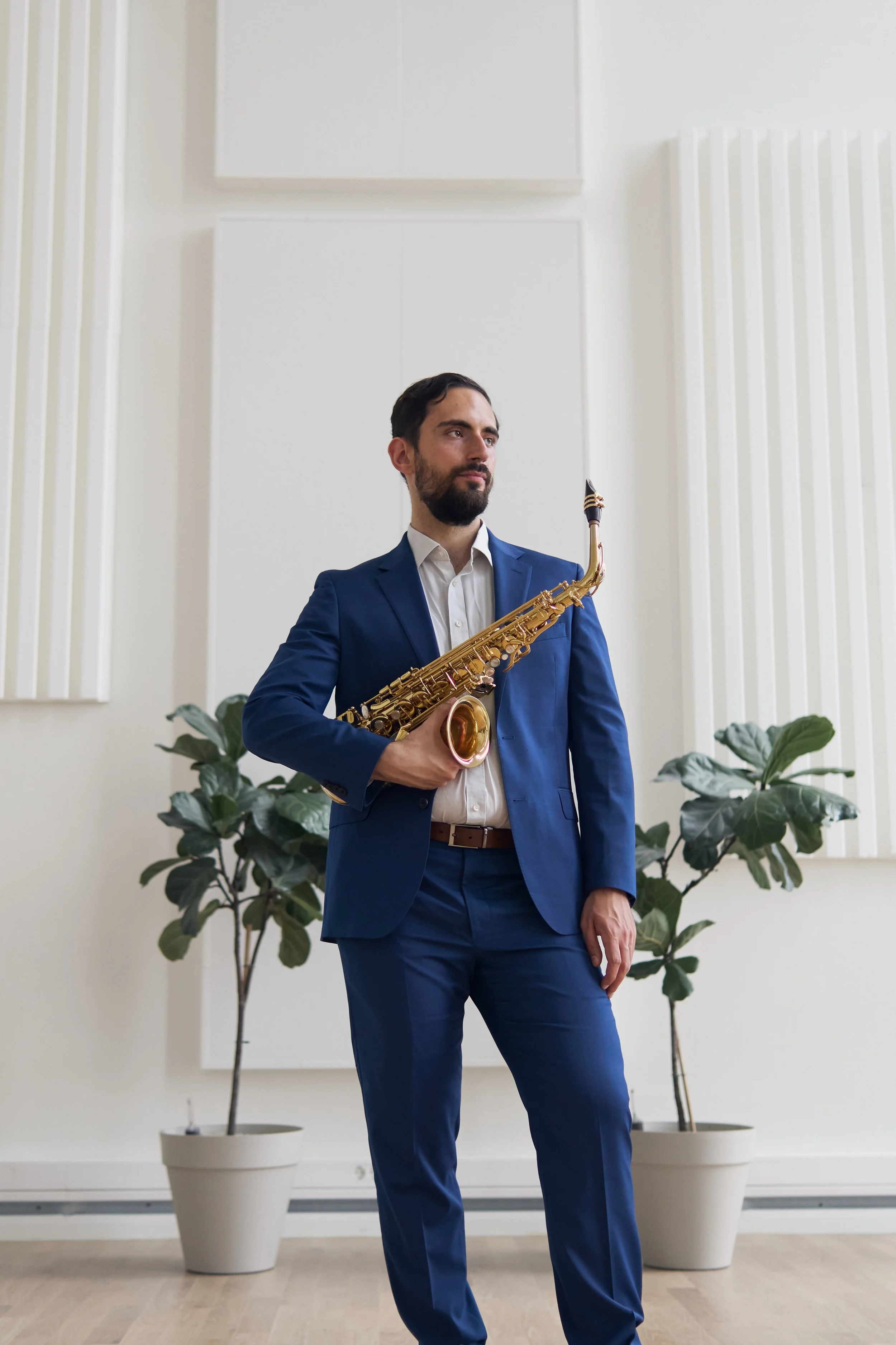 Man with dark hair and beard in a blue suit holding a saxophone stands in front of a white wall with potted plants.